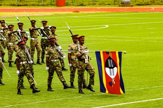 Group of Swazi soldiers in uniform participating in a ceremonial march with a national flag.