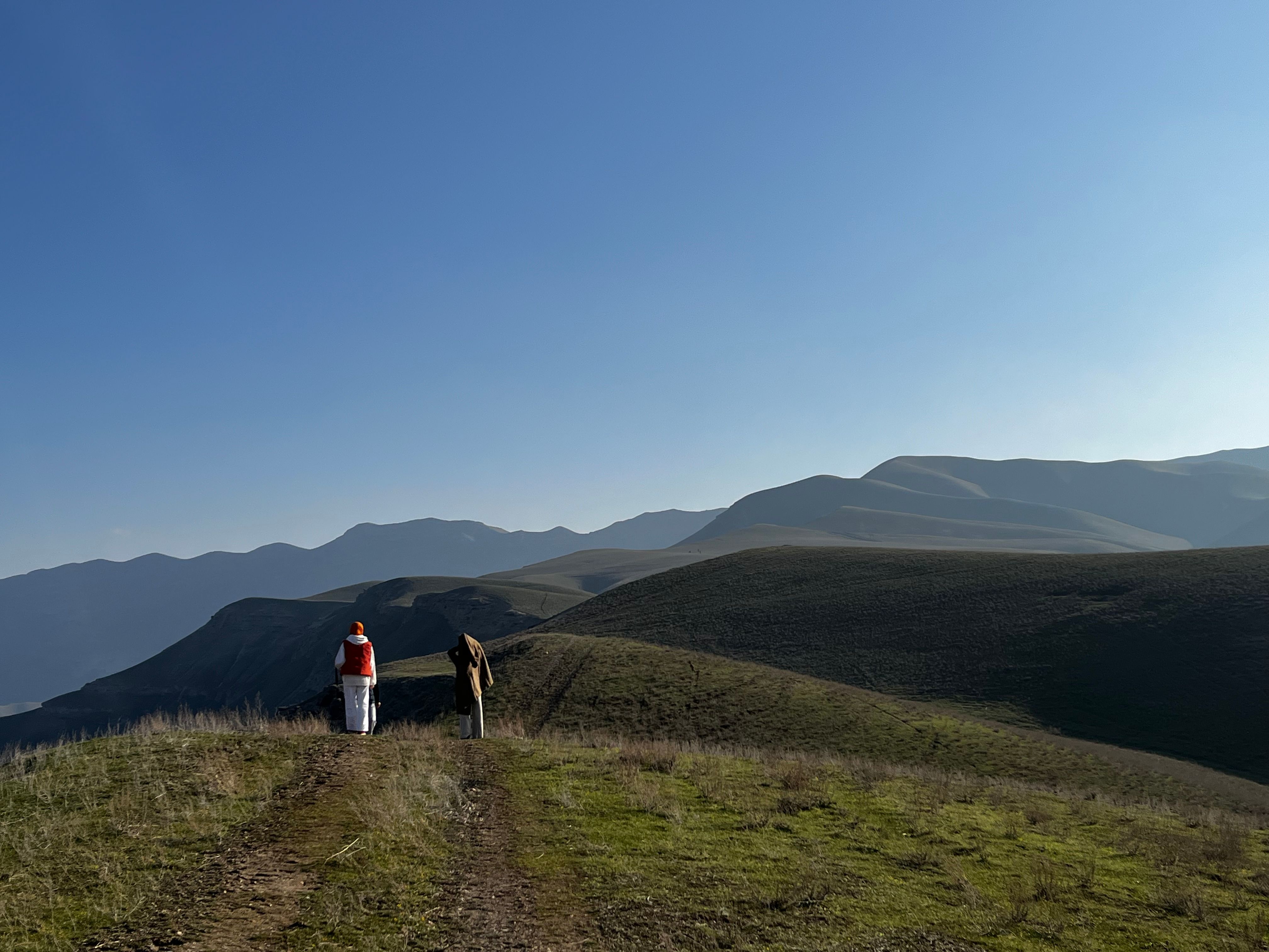 Two hikers explore a vast, rolling mountain landscape under a clear blue sky.