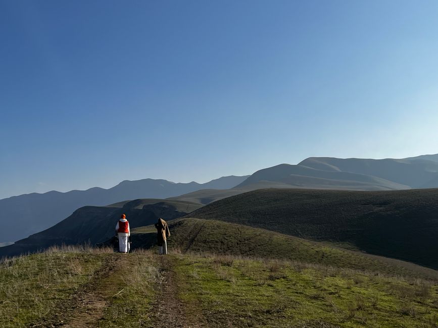 Two hikers explore a vast, rolling mountain landscape under a clear blue sky.