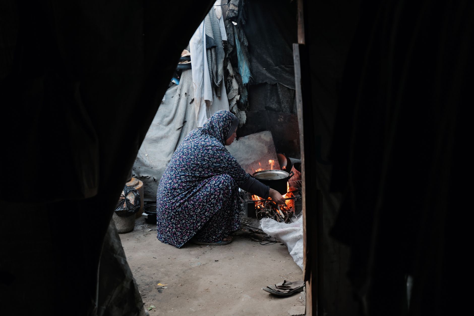 A woman in traditional attire kneeling while cooking over an open fire within a rustic shelter.