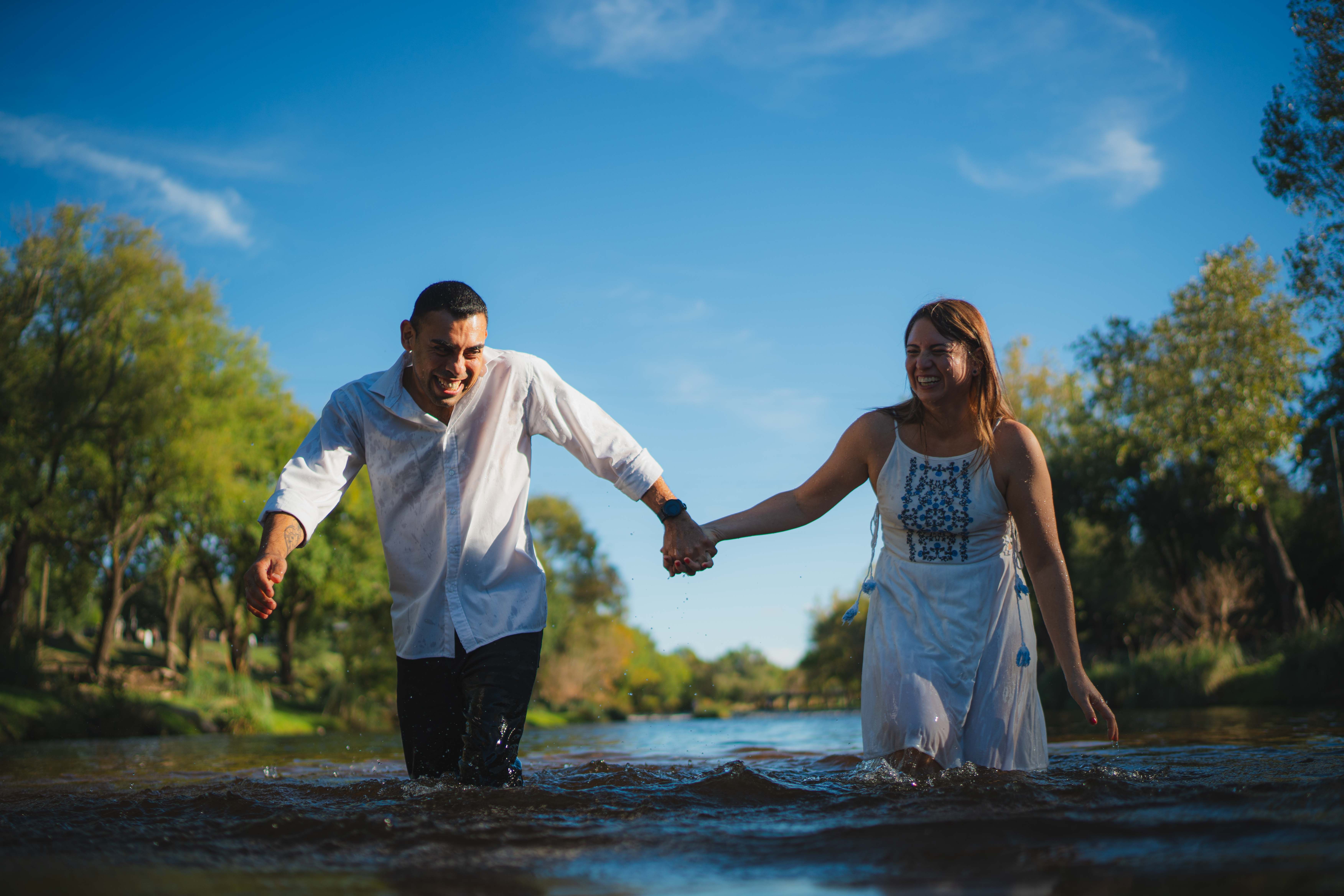 Smiling couple enjoying a playful moment as they walk hand-in-hand through a river under a vibrant blue sky.