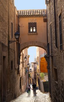 A couple walks under a brick arch in a historic alleyway in Toledo, Spain.