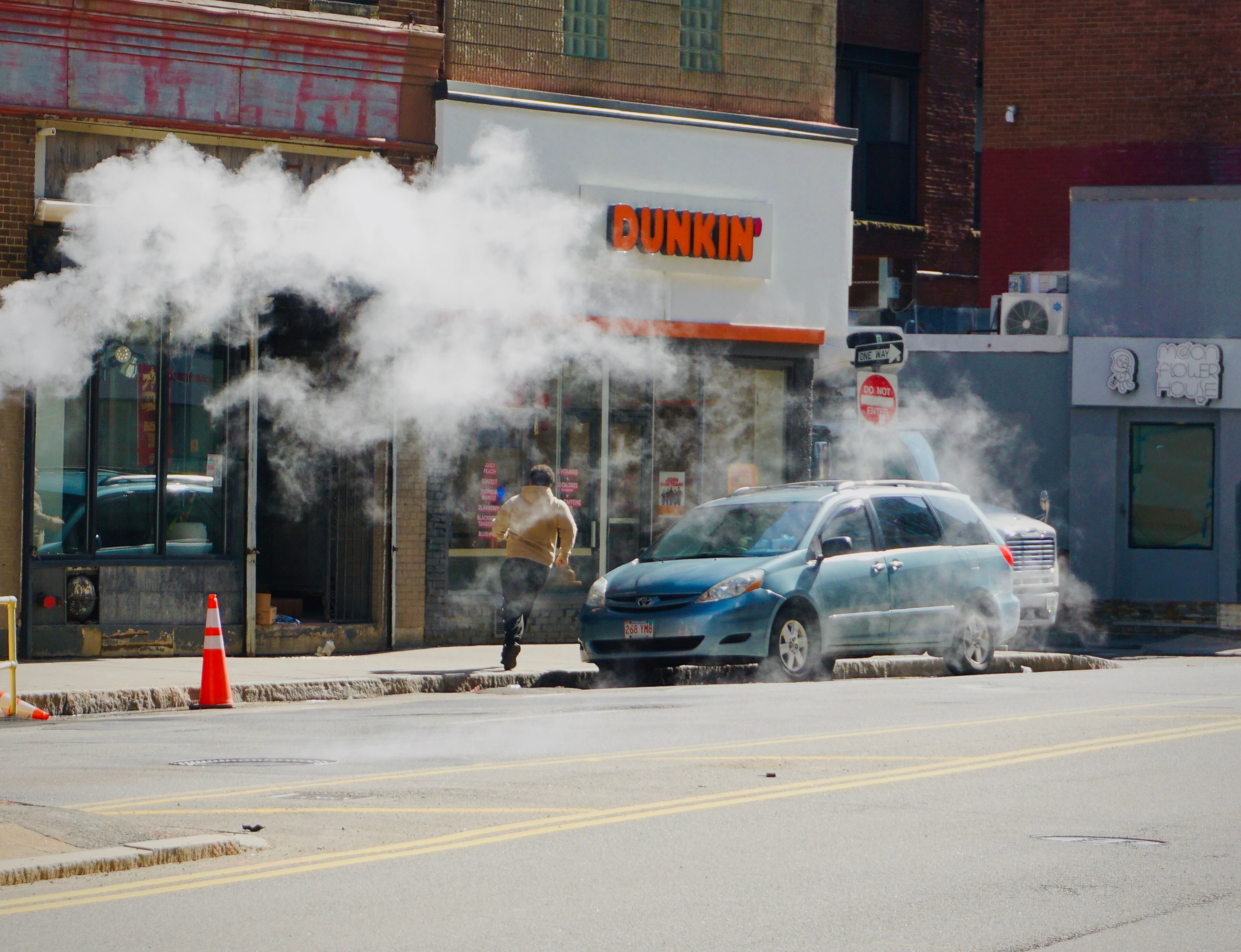 Urban street scene showing steam rising near a parked car by a Dunkin' shop, person walking away.