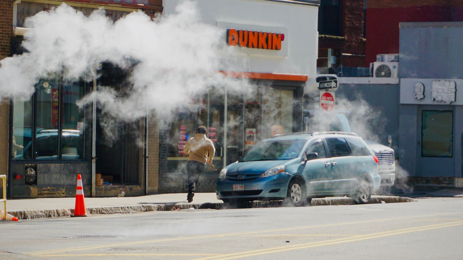 Urban street scene showing steam rising near a parked car by a Dunkin' shop, person walking away.