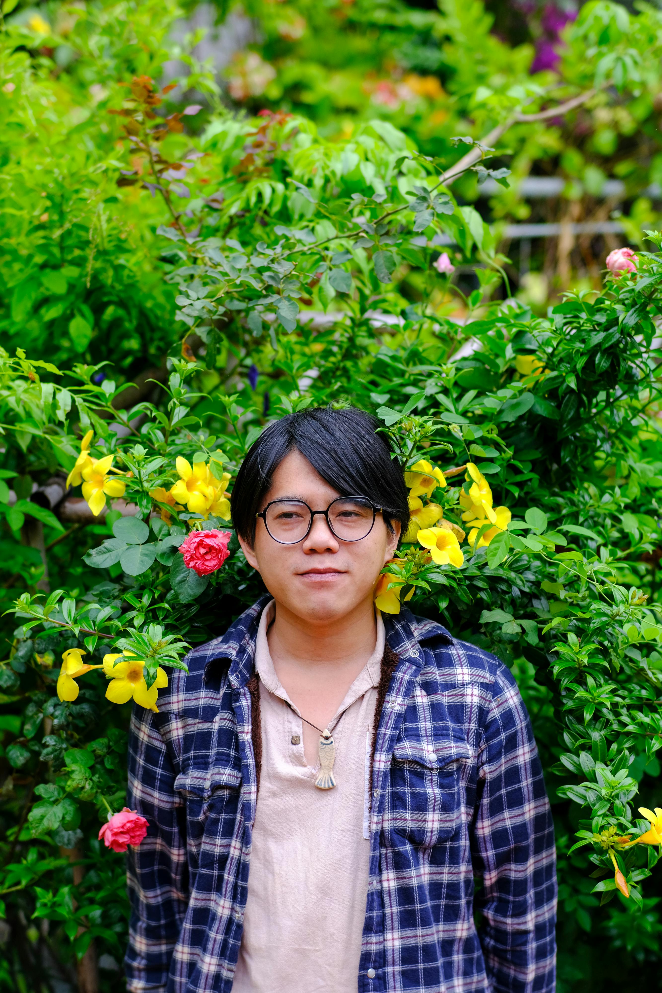 Young man in flannel poses in a lush garden in Hue, Vietnam, surrounded by vibrant flowers.
