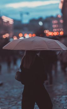 Woman with an umbrella walking on a rainy evening, city lights softly glowing in the background.