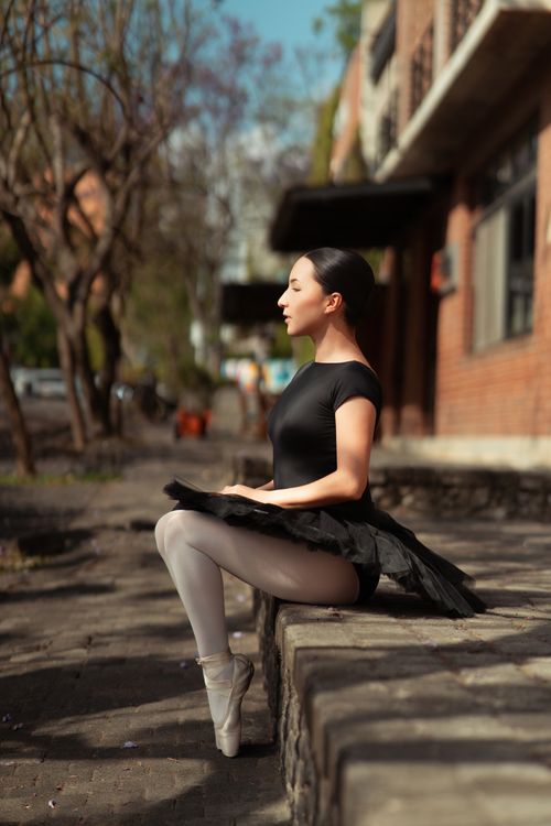 Free A ballerina in a black tutu rests on a stone wall outdoors on a sunny day. Stock Photo