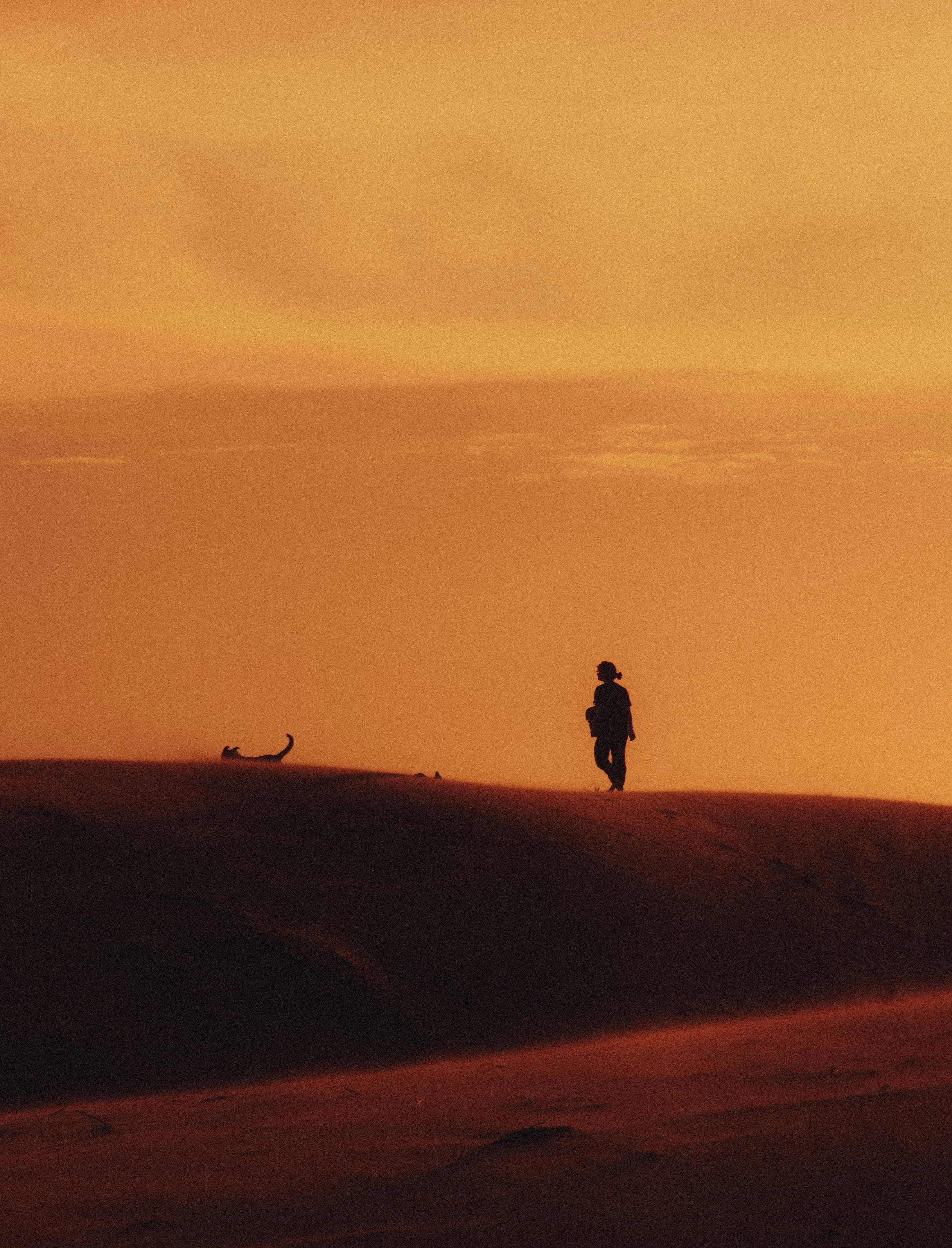 A lone figure traverses a sand dune under a vibrant orange sunset sky, symbolizing solitude and adventure.