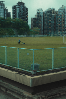 A person mowing a grass field in an urban park with tall buildings in Hong Kong.