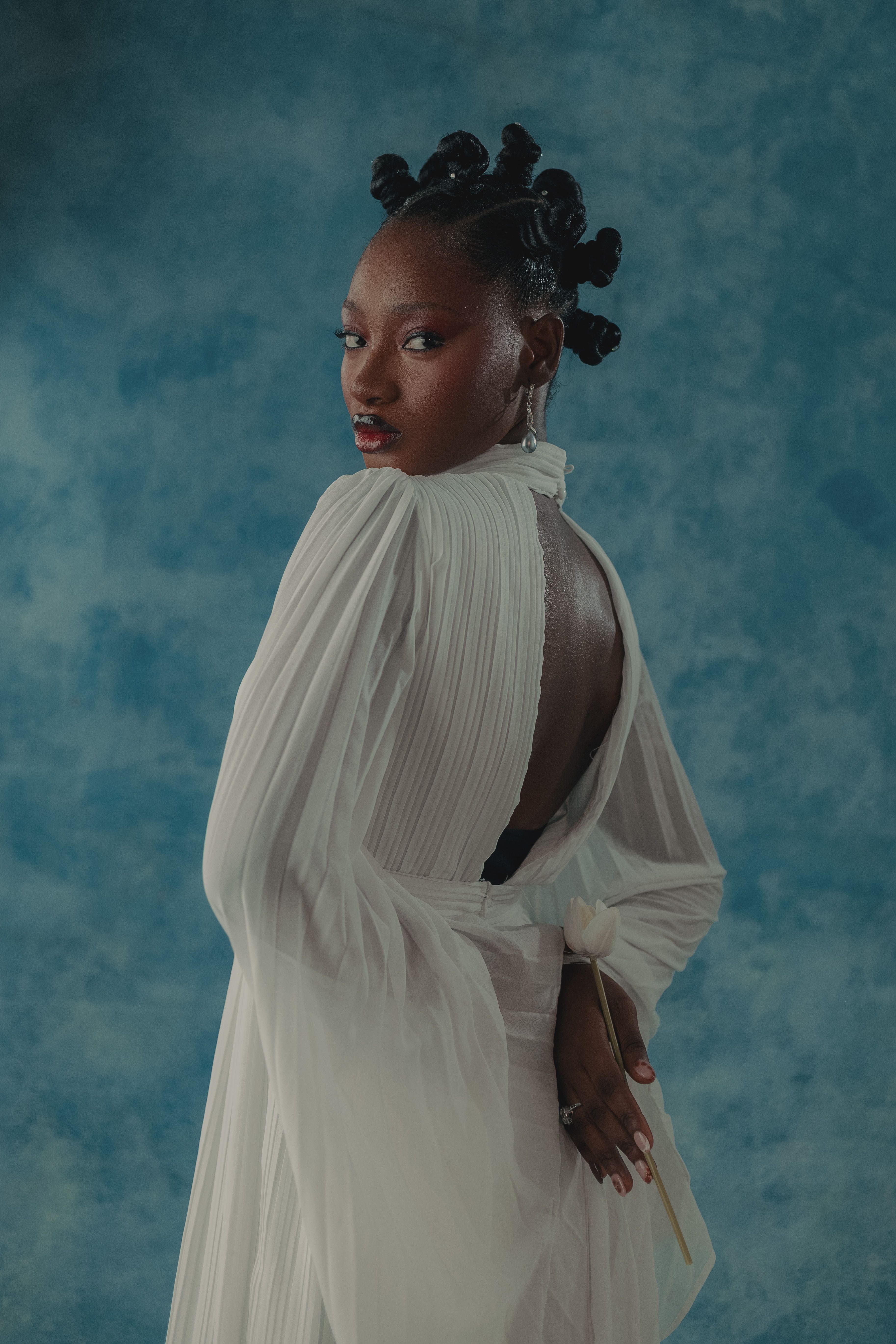 Elegant portrait of a woman in a pleated white dress with intricate braided hairstyle against a blue background.