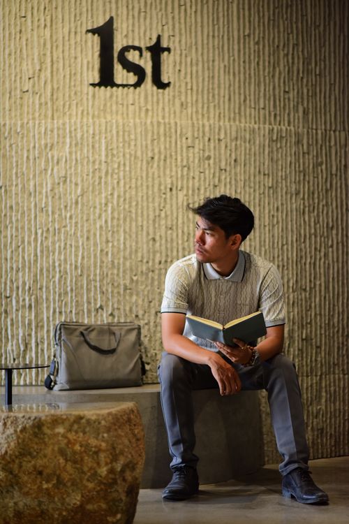 Free Adult male reading in a modern interior, sitting against textured wall with '1st'. Stock Photo