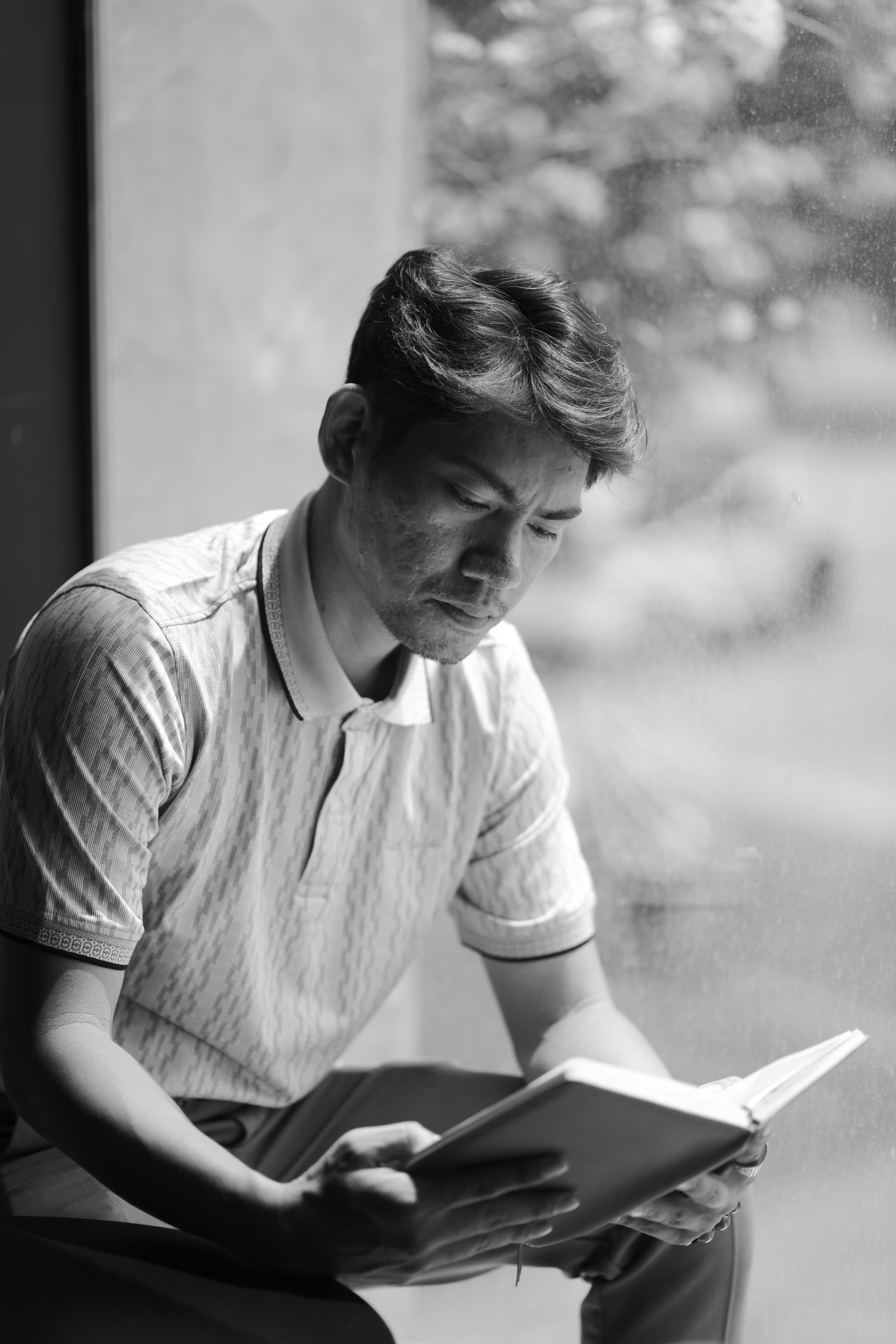 Black and white portrait of a thoughtful man reading a book by a window.
