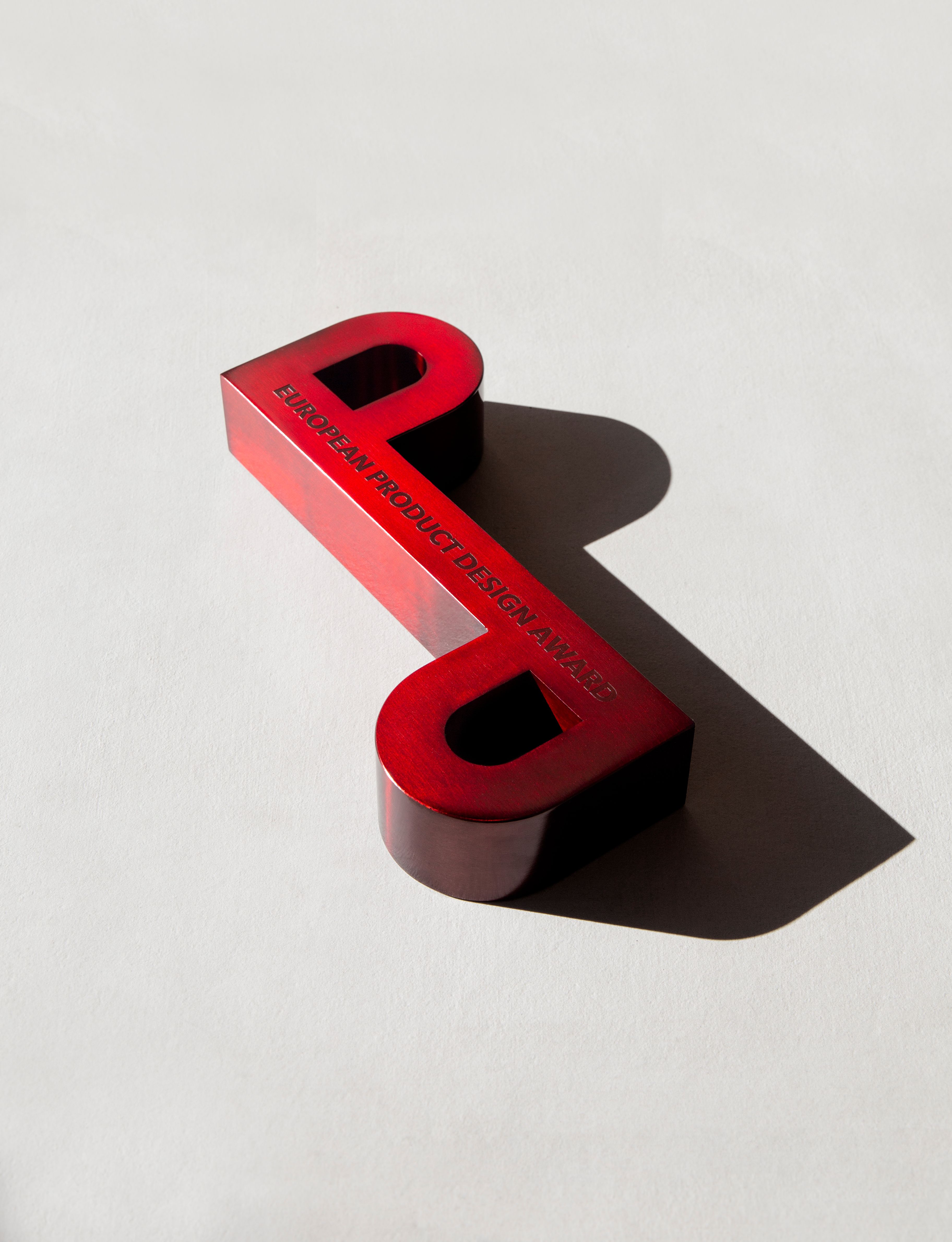 Red trophy shaped as 'D/P' casting a shadow on a plain surface, symbolizing design excellence.