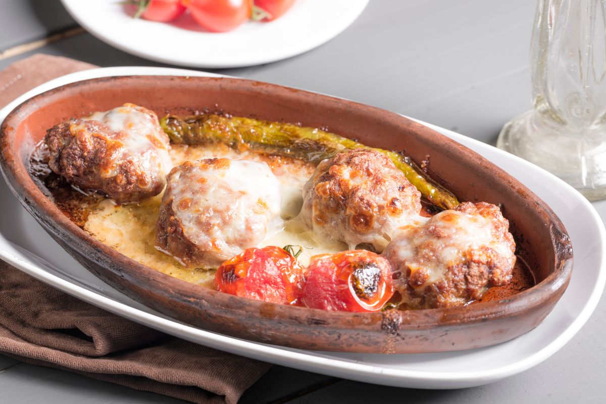Clay baking dish of browned meatballs under melted cheese with roasted tomatoes and green pepper
