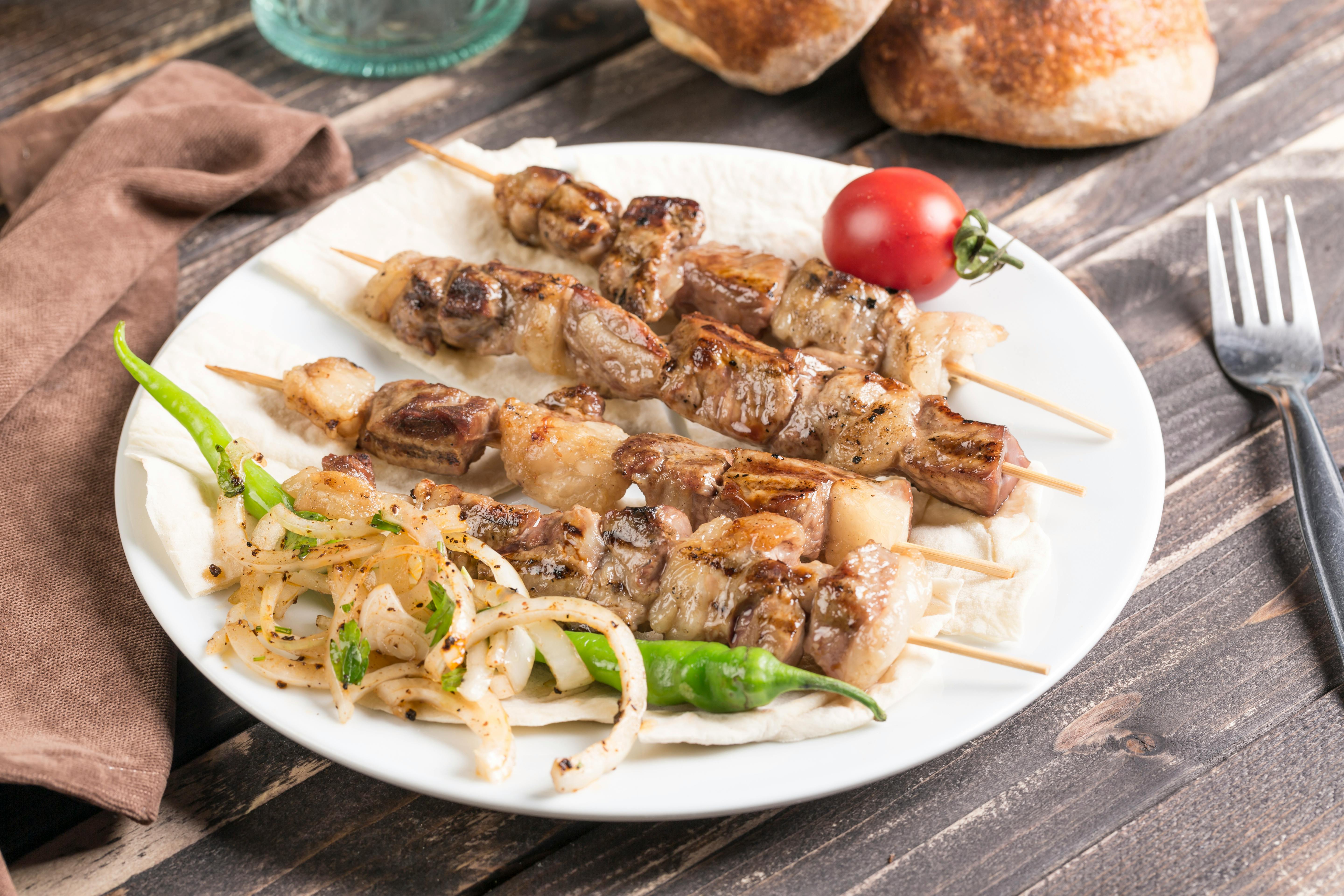 Charred lamb souvlaki on metal skewers served on a white plate with a small bowl of chunky white tzatziki, warm pita bread, and sliced red onions