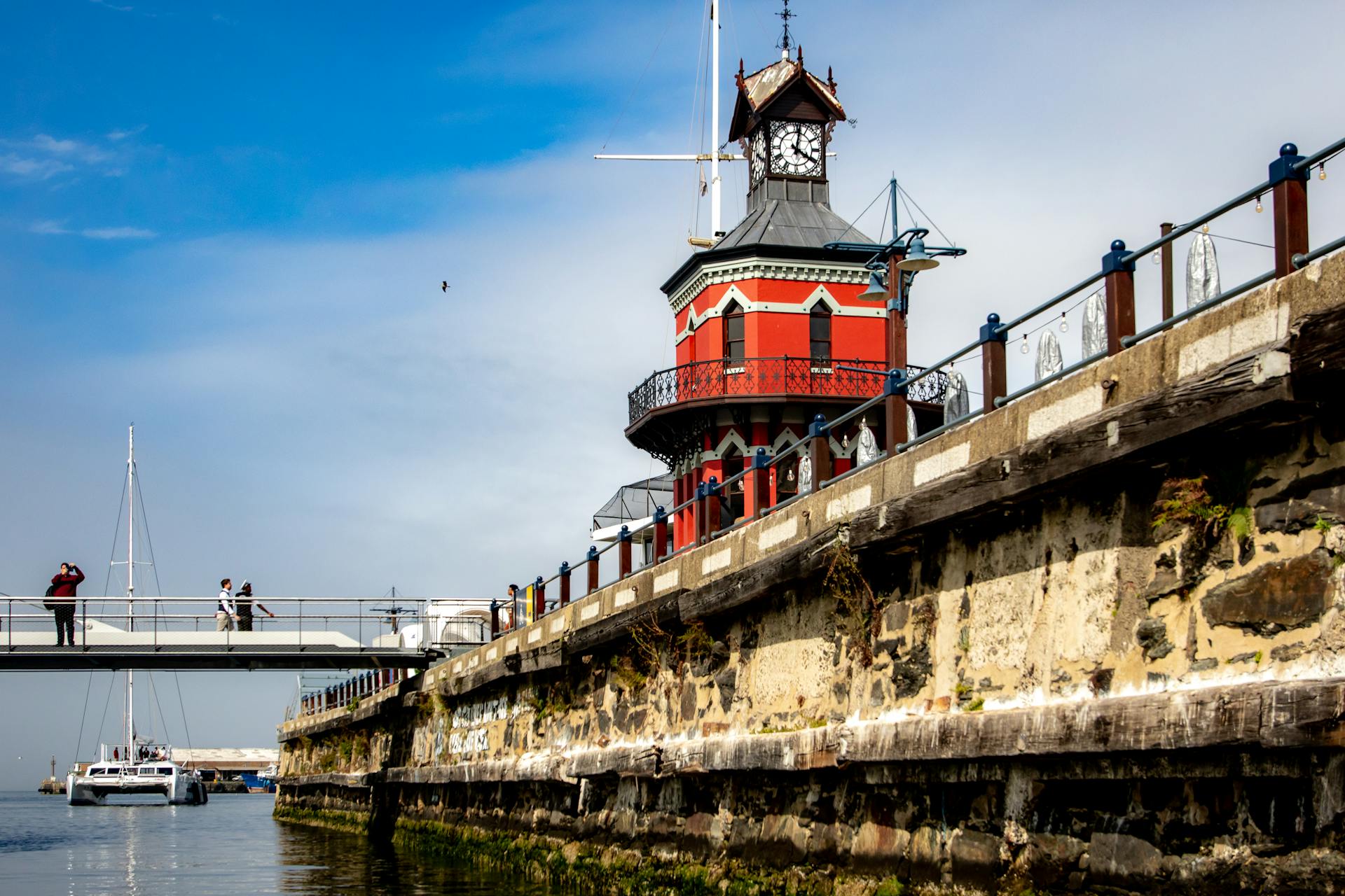 Scenic view of a historic clock tower at the waterfront in Cape Town, featuring a bridge and passing yacht.
