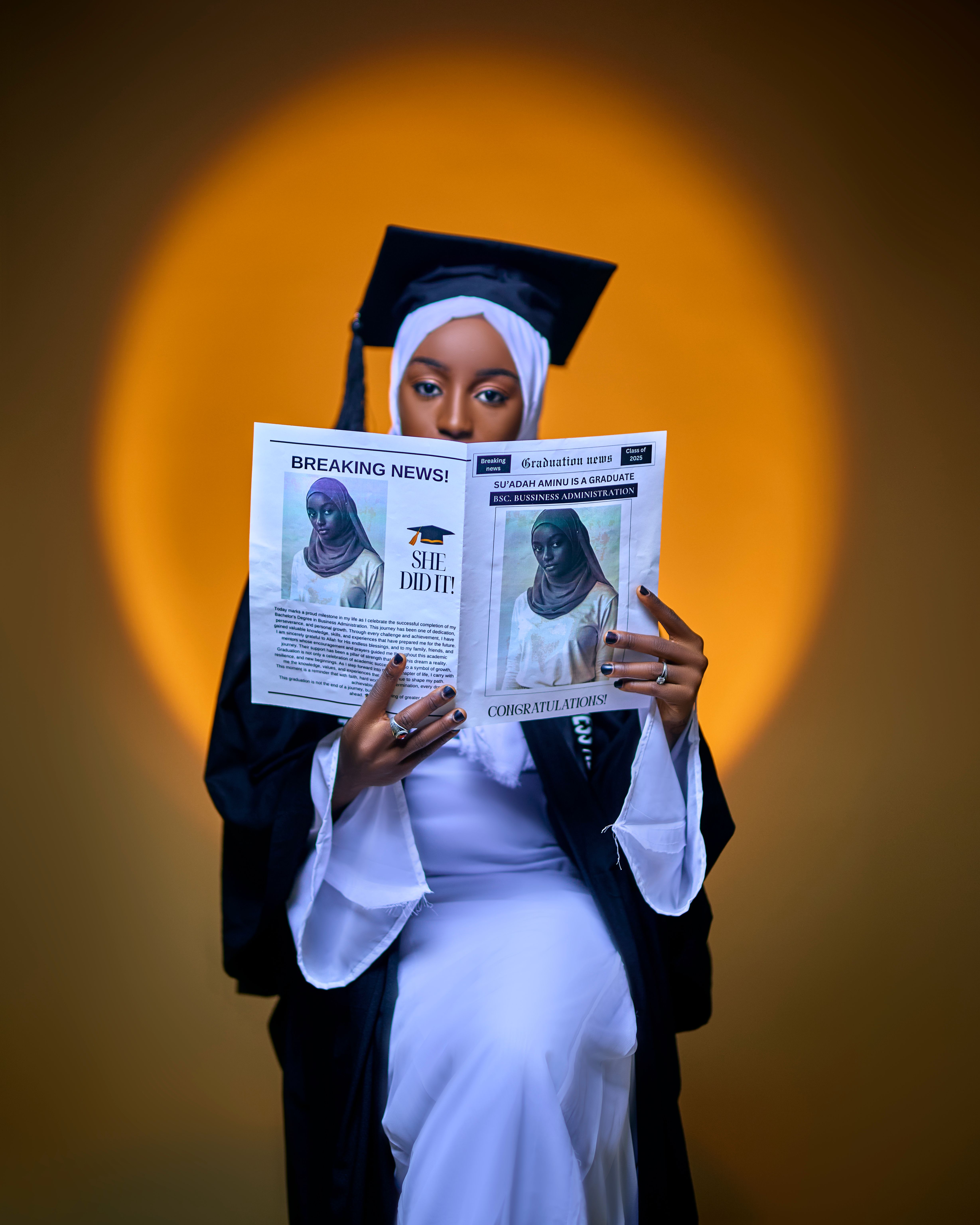 A female graduate in cap and gown joyfully reads a congratulatory newspaper.