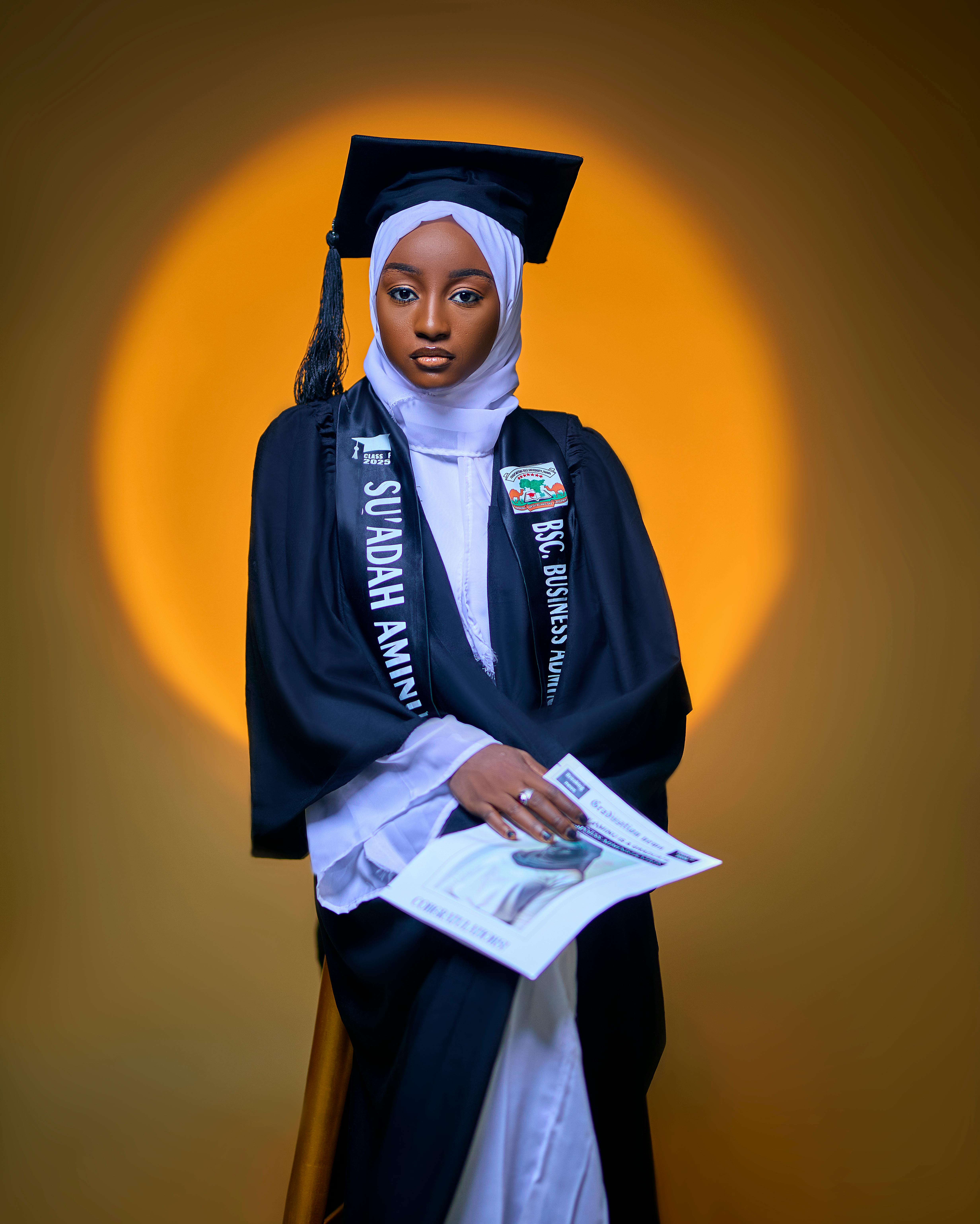 A young woman in graduation attire holding a certificate, posed against a warm backdrop.