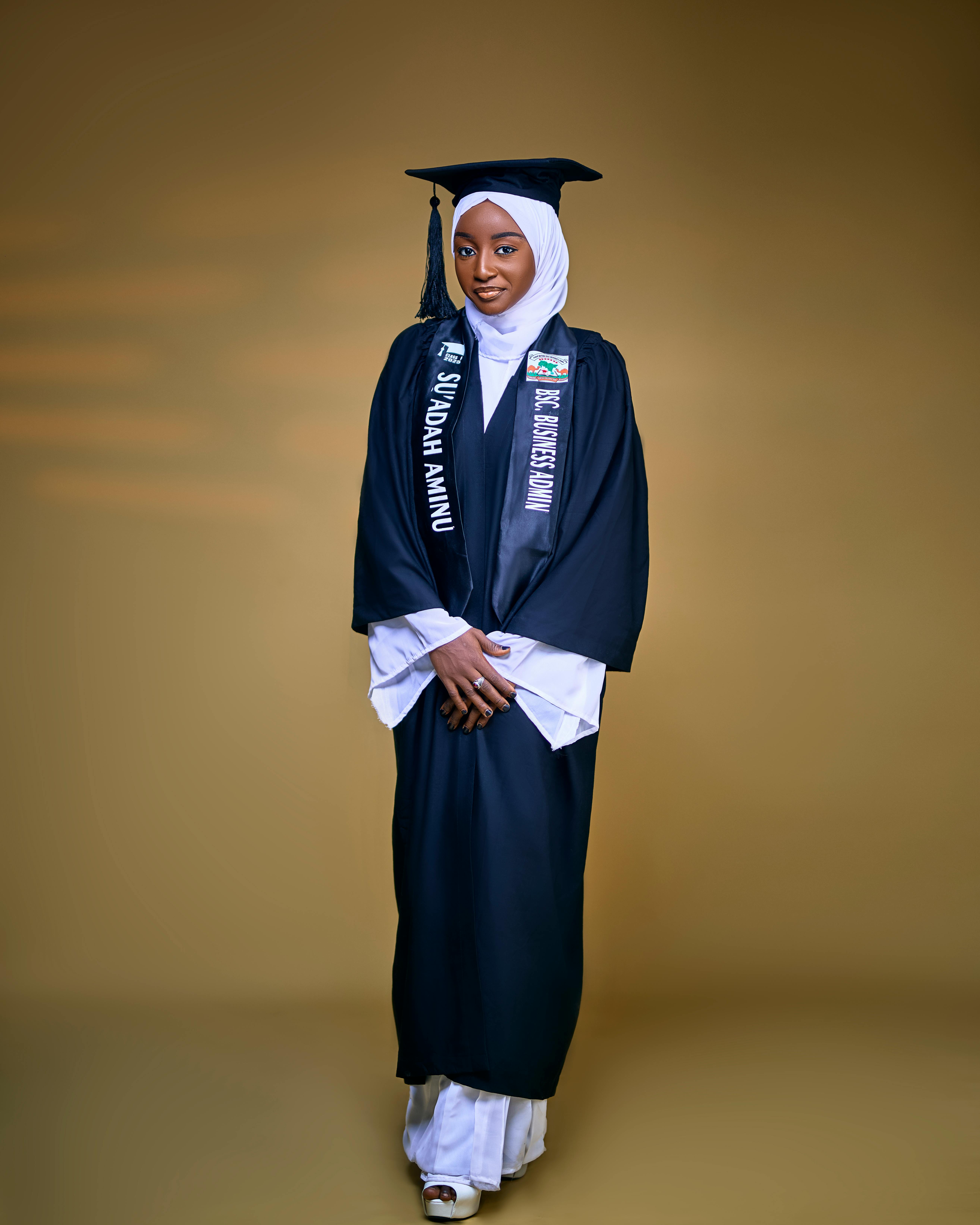 Young woman in graduation attire with hijab on golden background, symbolizing achievement.