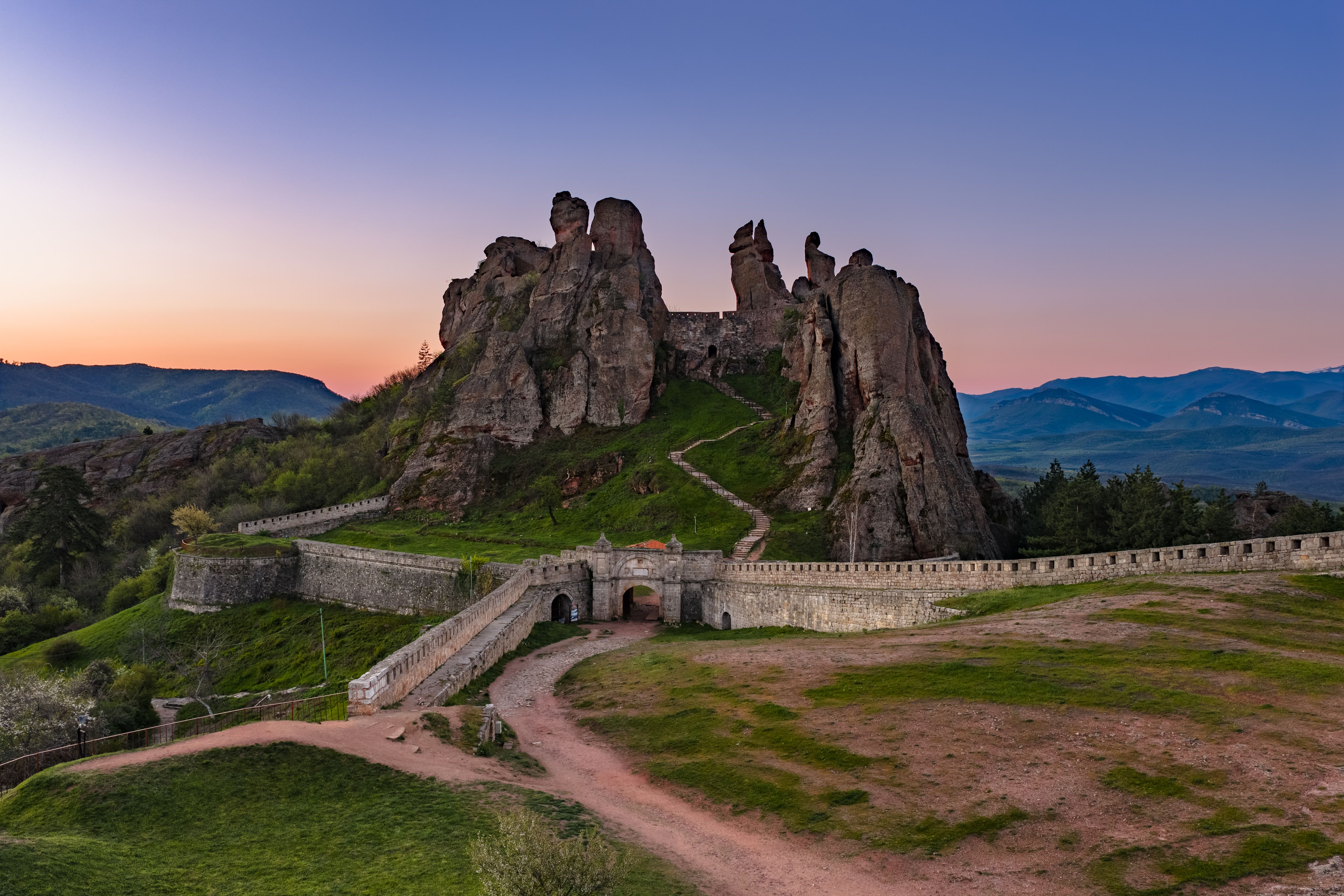 Belogradchik Rocks at sunset with dramatic sandstone formations glowing in warm light, lush greenery in foreground