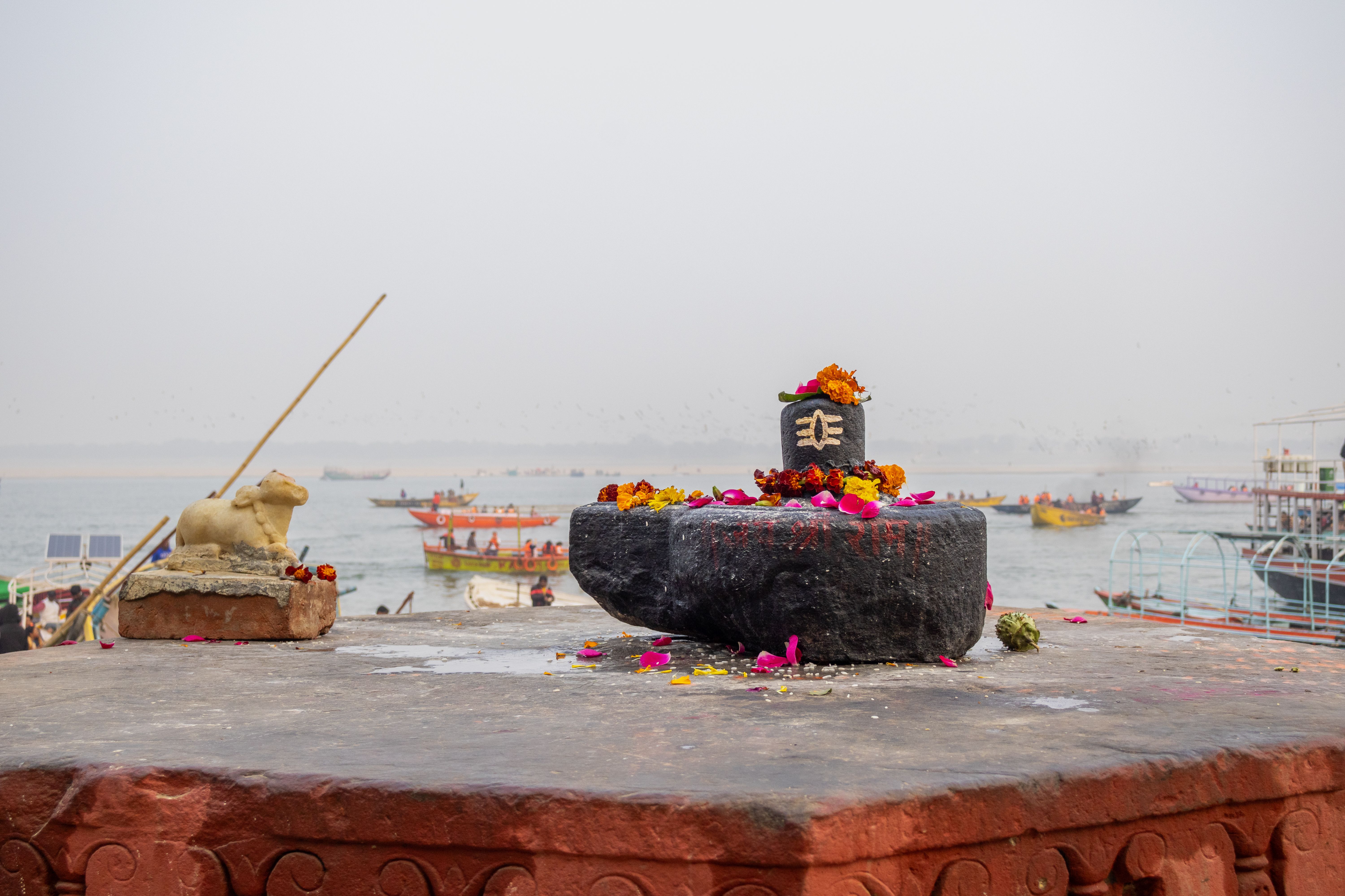 Sacred Shiva Lingam adorned with flowers by the Ganges River, India, with colorful boats in the background.