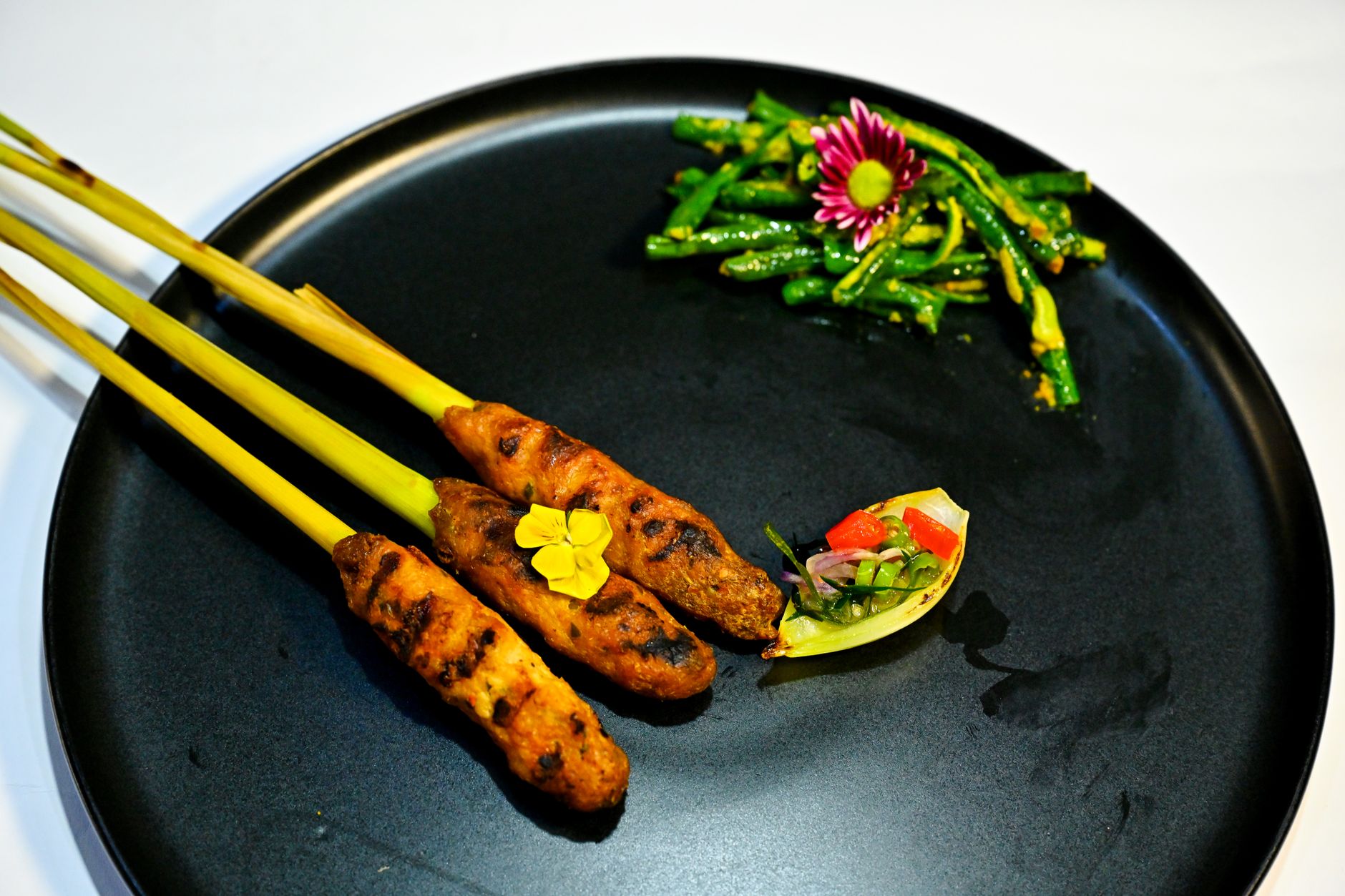 Close-up of Sate Lilit skewers on a black plate with vibrant side dishes.
