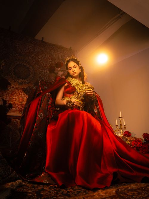 Free A woman in a red gown with gold accents sits elegantly under warm ambient lighting, evoking a regal atmosphere. Stock Photo