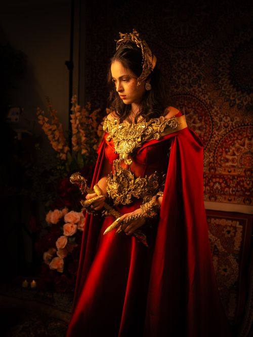 Free A woman in a regal red gown and gold accessories, set against a tapestry backdrop. Stock Photo
