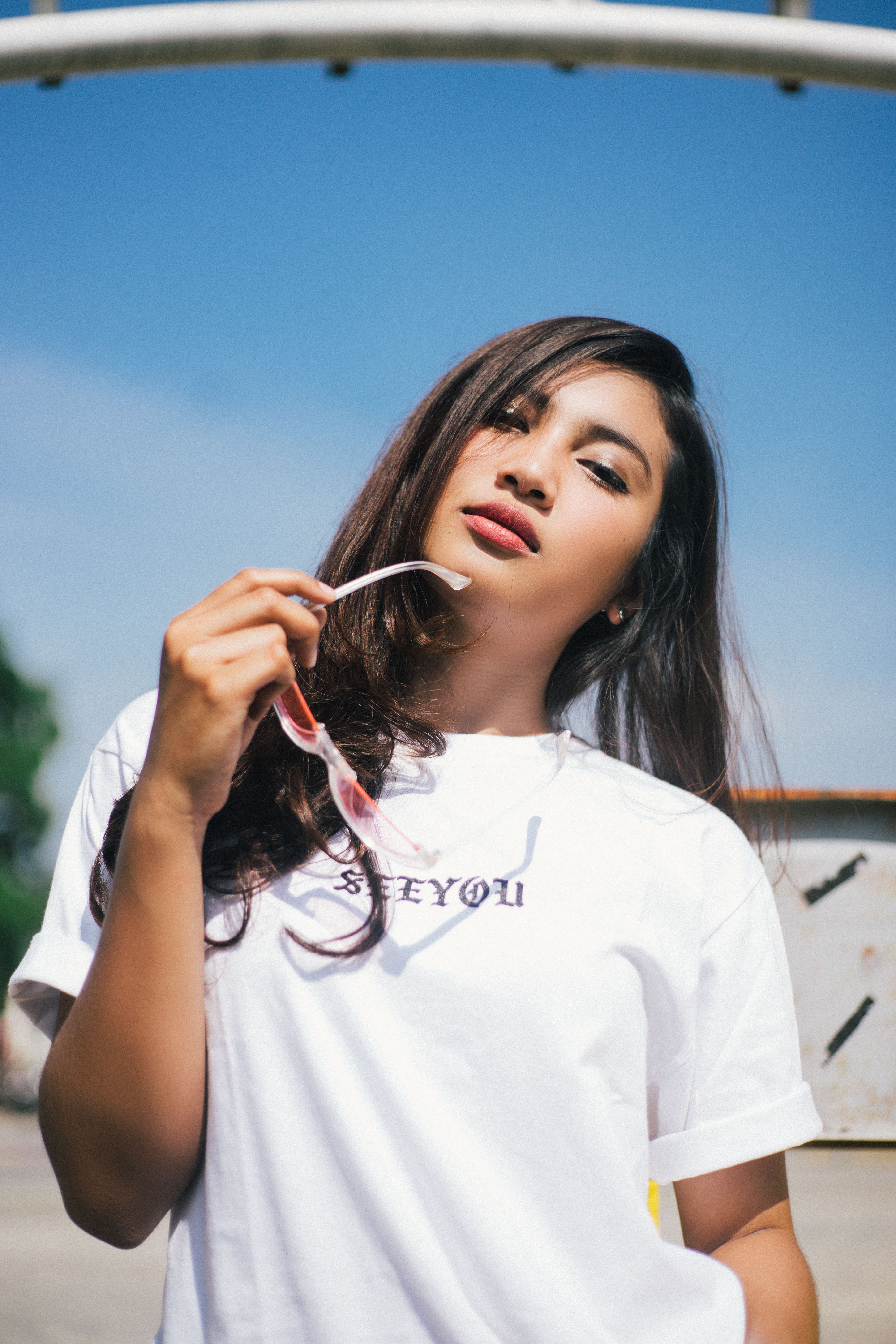 Brunette woman posing with sunglasses outdoors, exuding confidence under the bright sky.