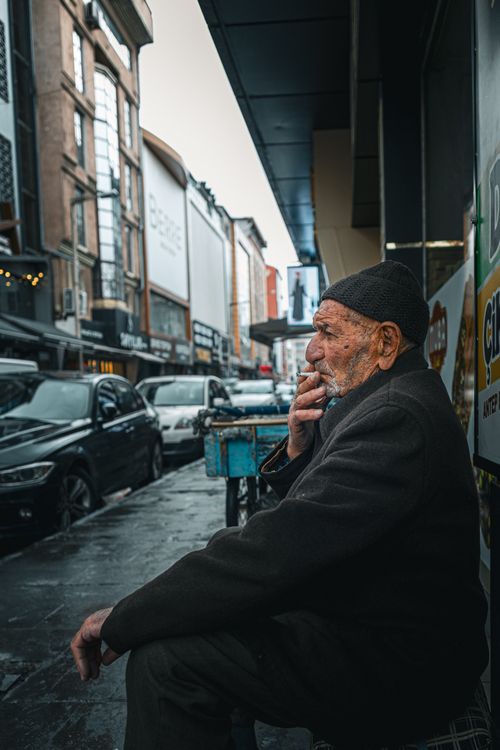 Free Elderly man sits pensively on a bustling city street, amidst urban architecture. Stock Photo