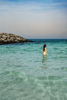 A young girl stands in the calm sea near rocky shore under a clear sky, embodying tranquility.
