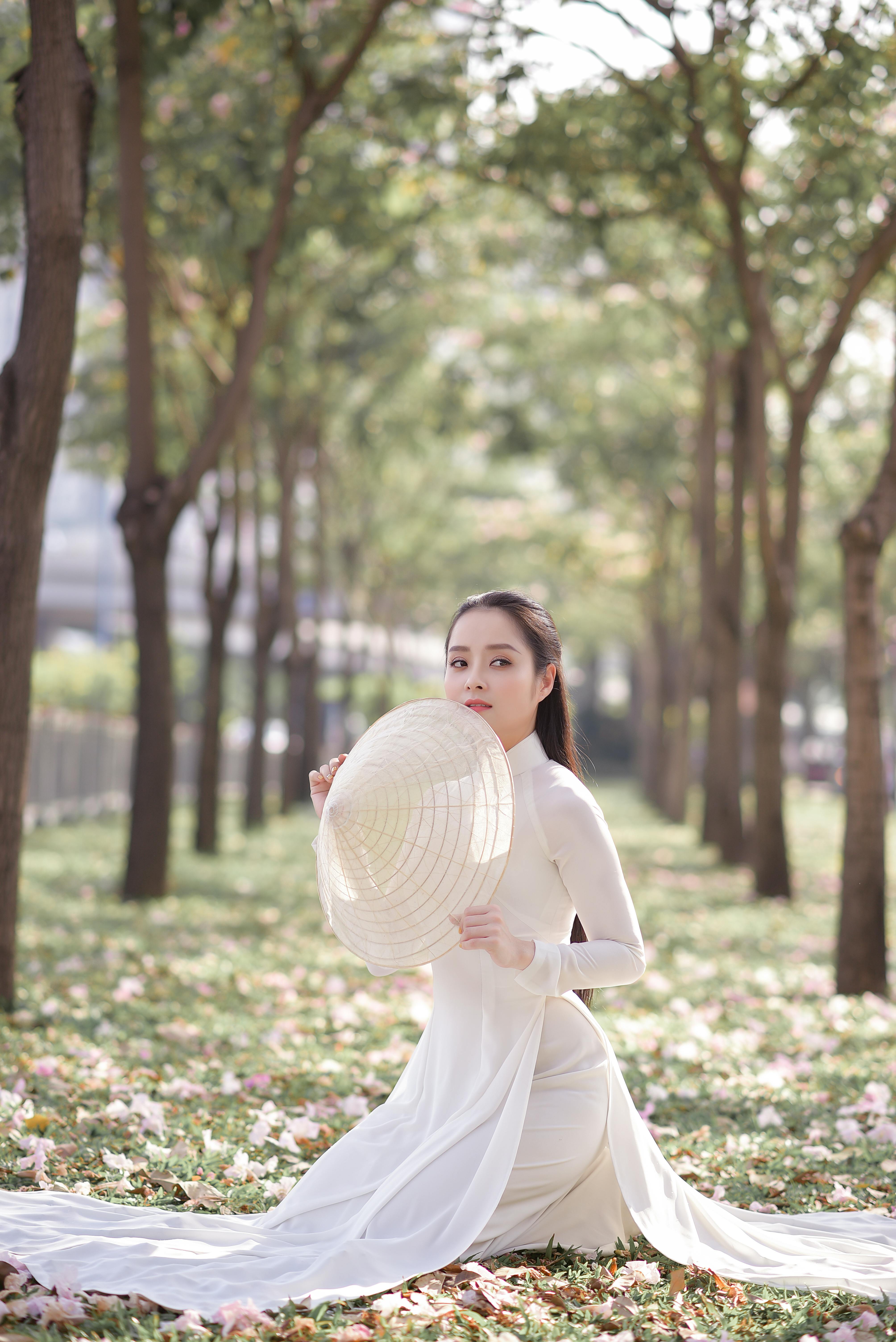 一位头戴草帽、身穿白色长裙的女子，坐在绿树成荫的小路上，显得优雅而宁静