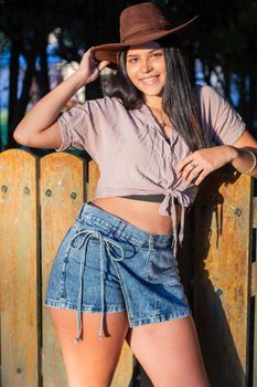 A young woman smiling confidently outdoors, wearing a brown hat and denim shorts.