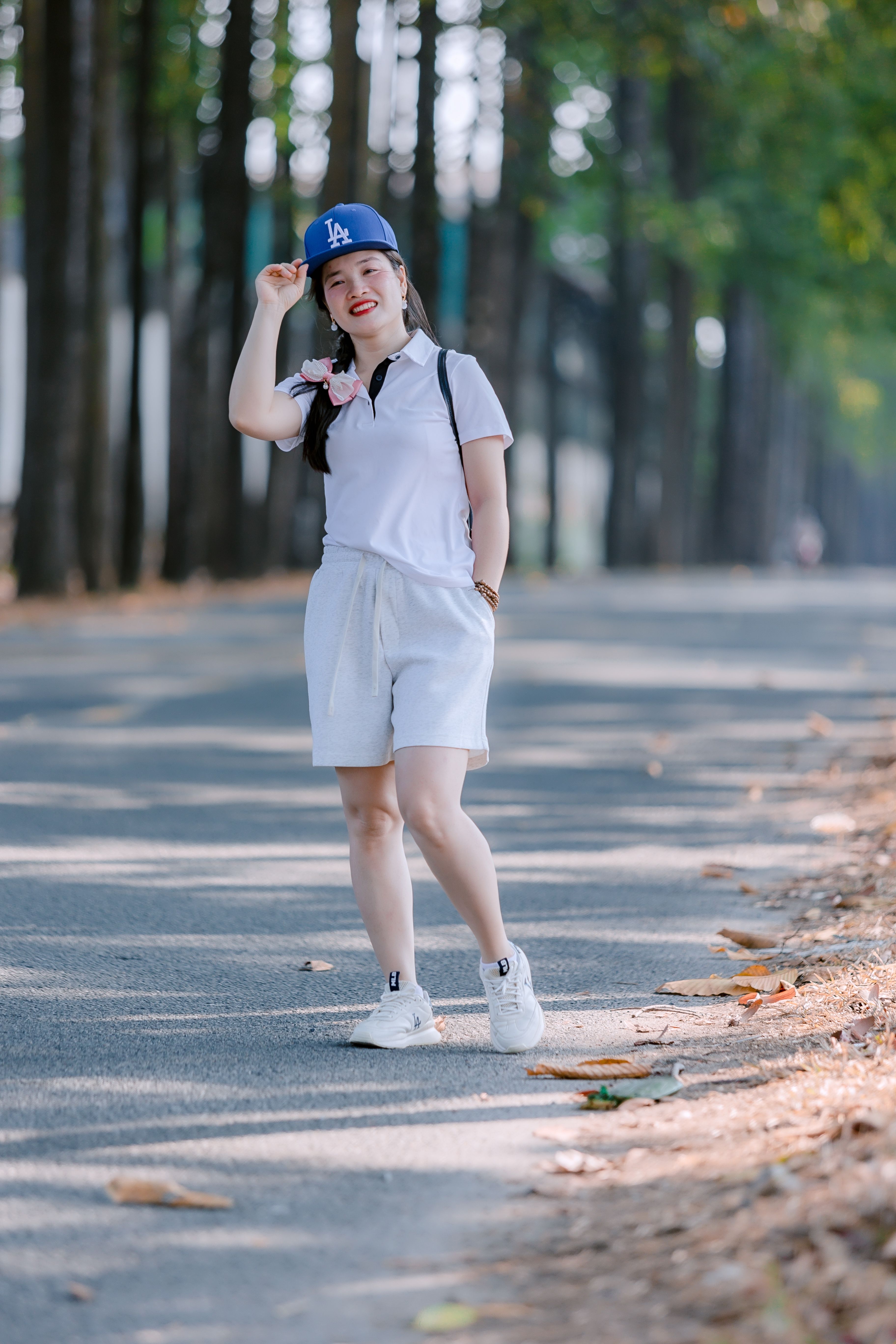 Smiling woman in white attire wearing a blue cap walking on a tree-lined path.
