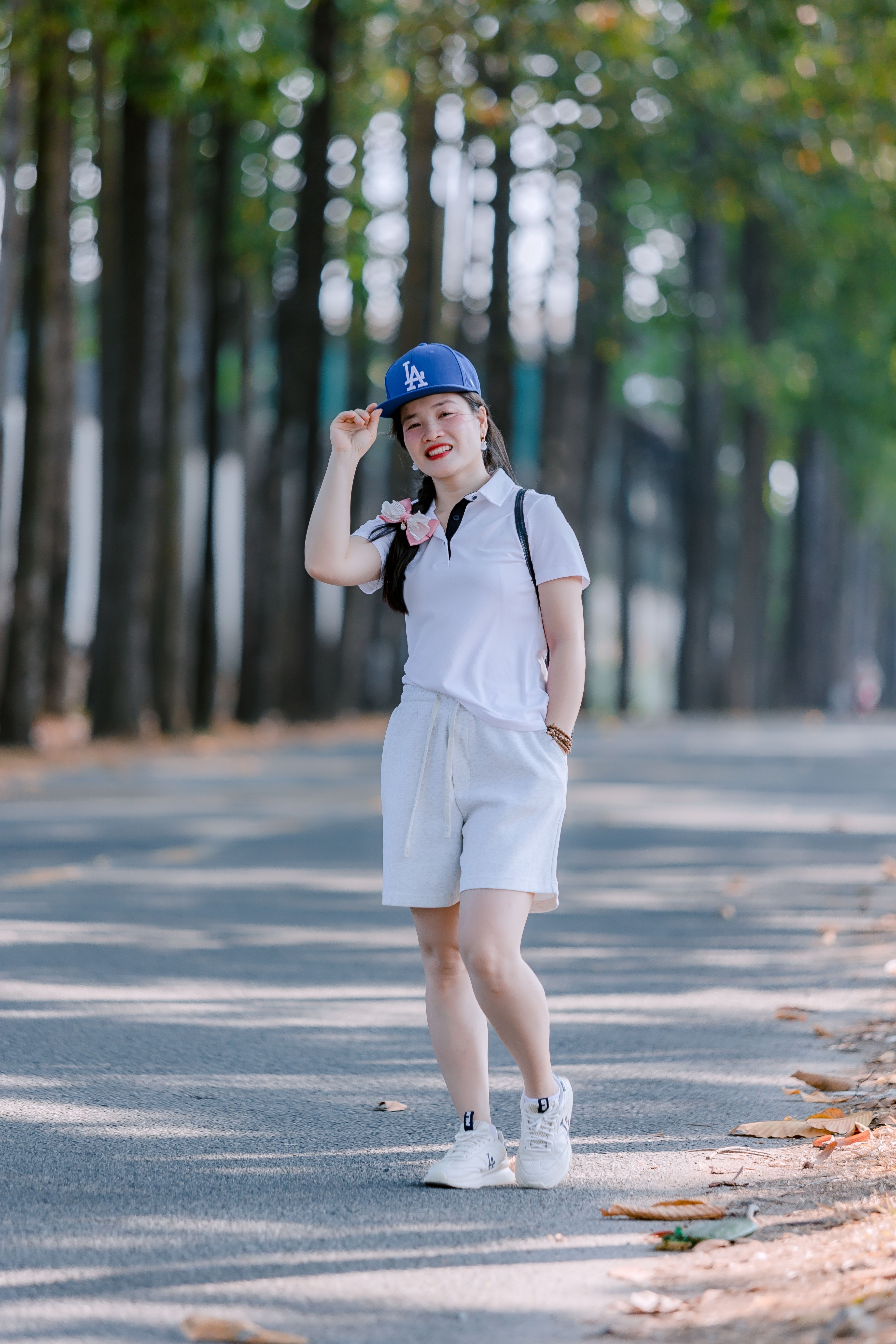 Woman in a white outfit and blue cap posing outdoors on a sunlit tree-lined avenue.