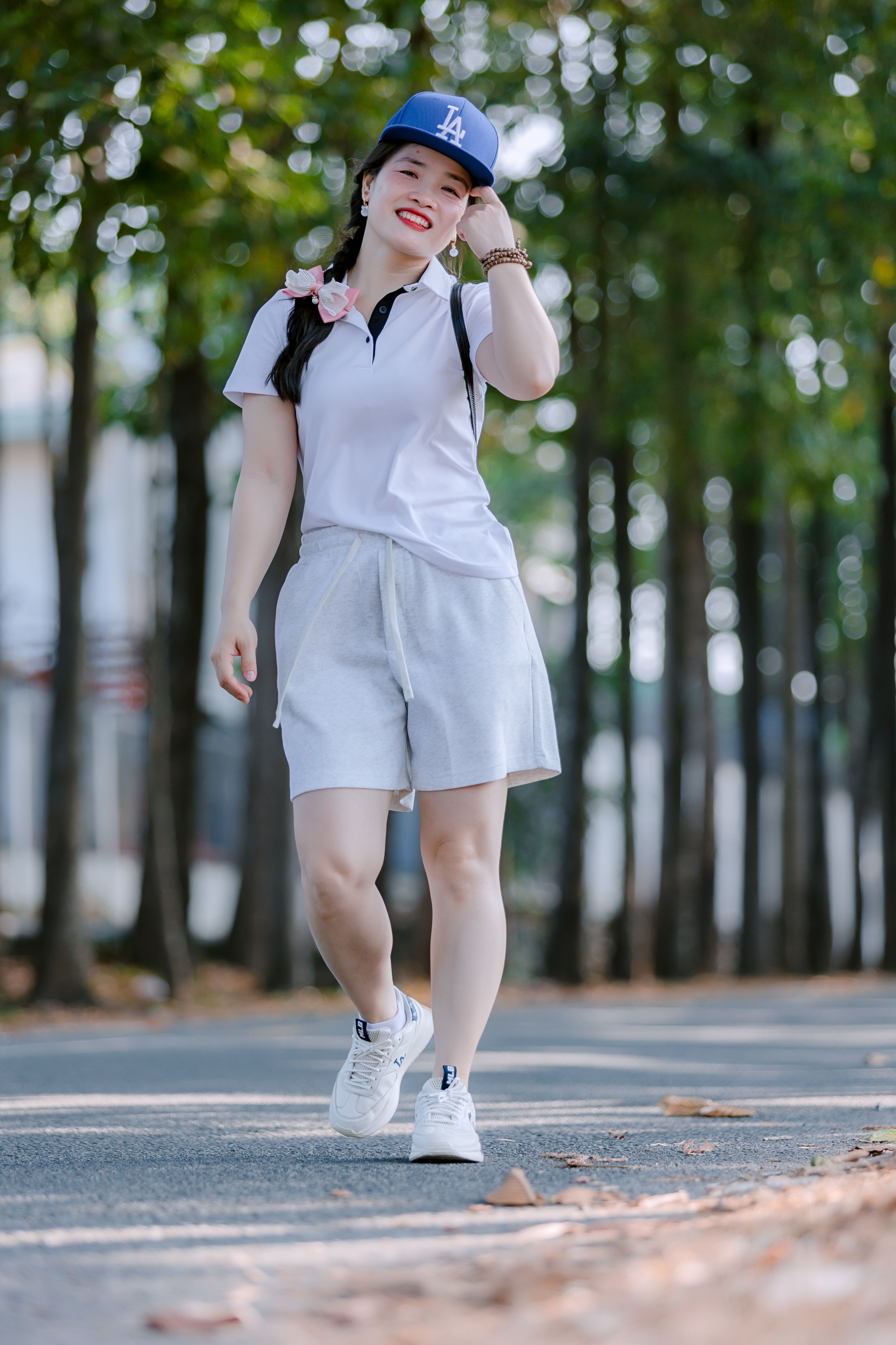 Smiling young woman in casual attire walking on a tree-lined path on a sunny day.