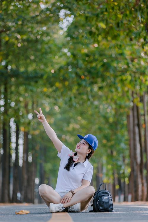 Free A woman sits on a tree-lined road enjoying nature, showing a peace sign. Stock Photo