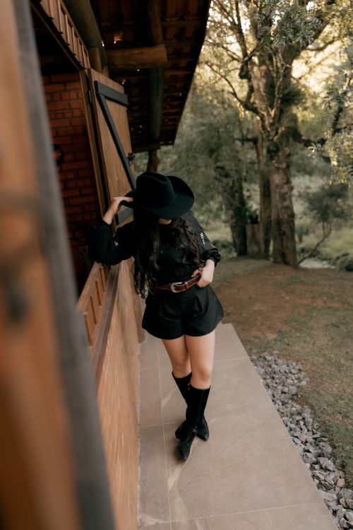 Free A stylish woman in cowboy attire poses by a cabin in a serene outdoor setting. Stock Photo