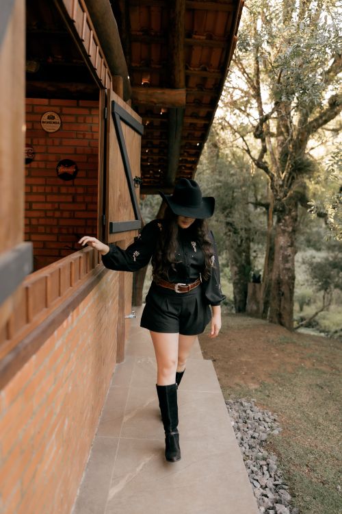 Free A woman wearing a cowboy hat and boots walks by a rustic brick house in a scenic outdoor setting. Stock Photo