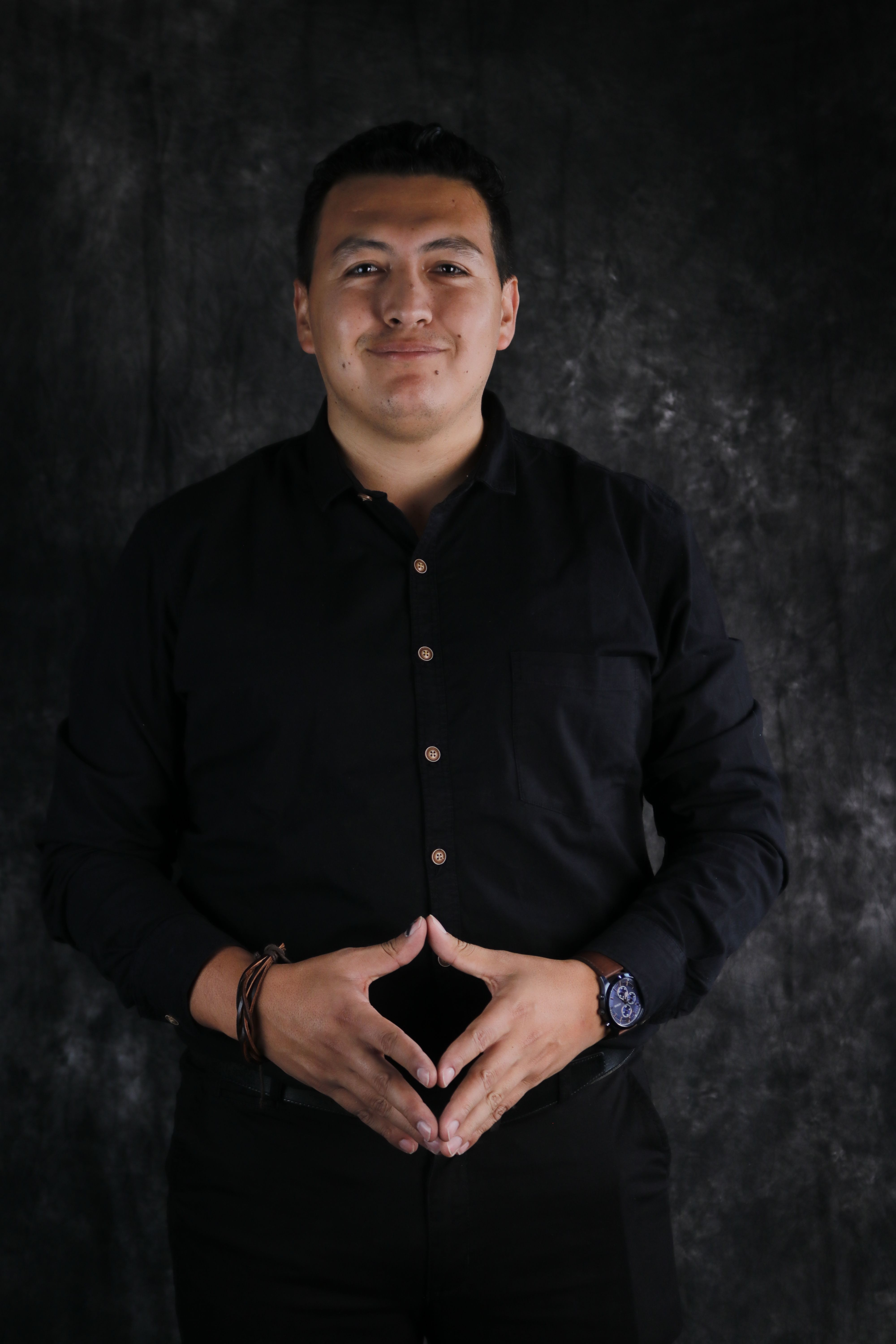 Smiling man in black shirt posing with hands clasped against dark background.