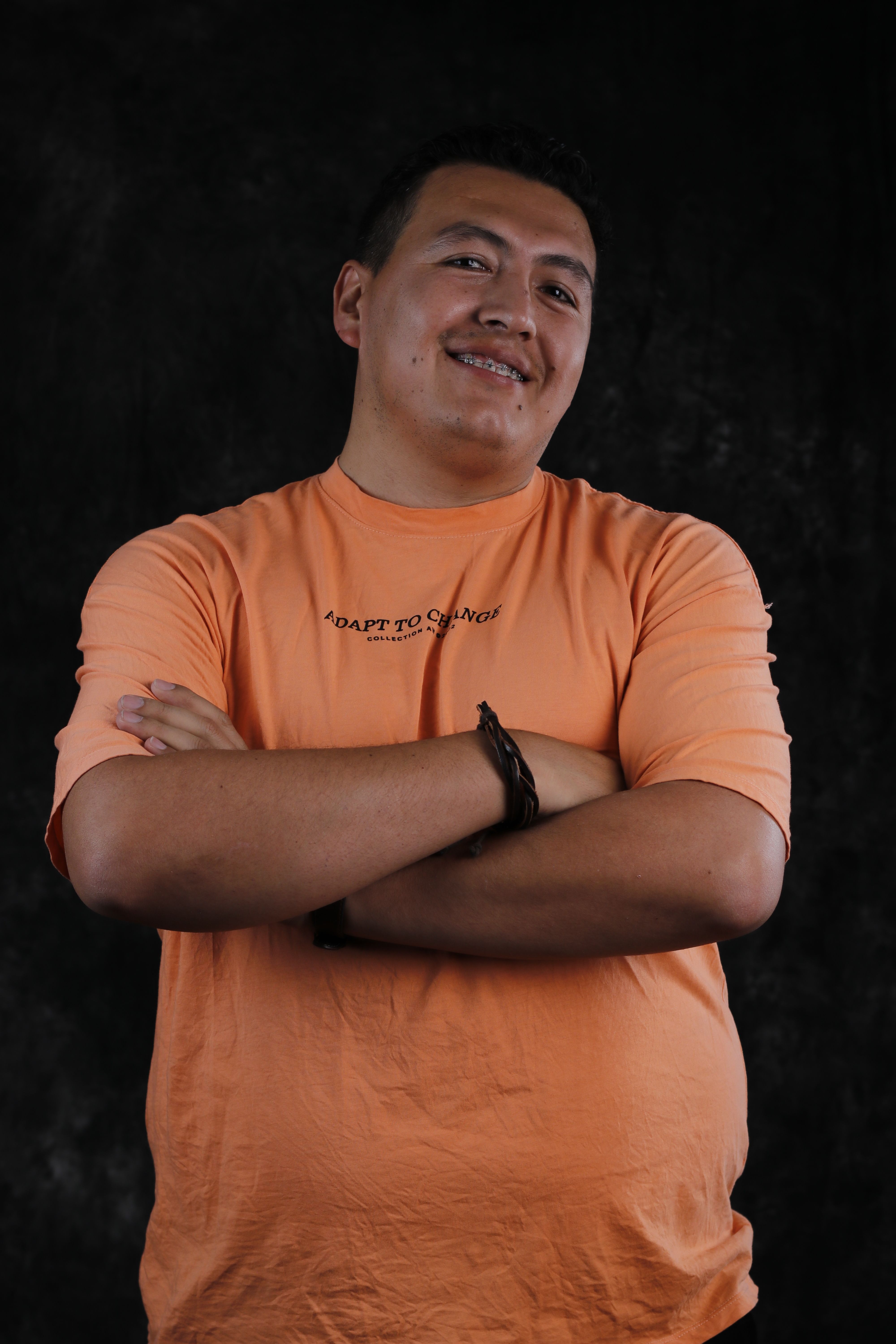 Portrait of a smiling man wearing an orange t-shirt with folded arms against a dark background.