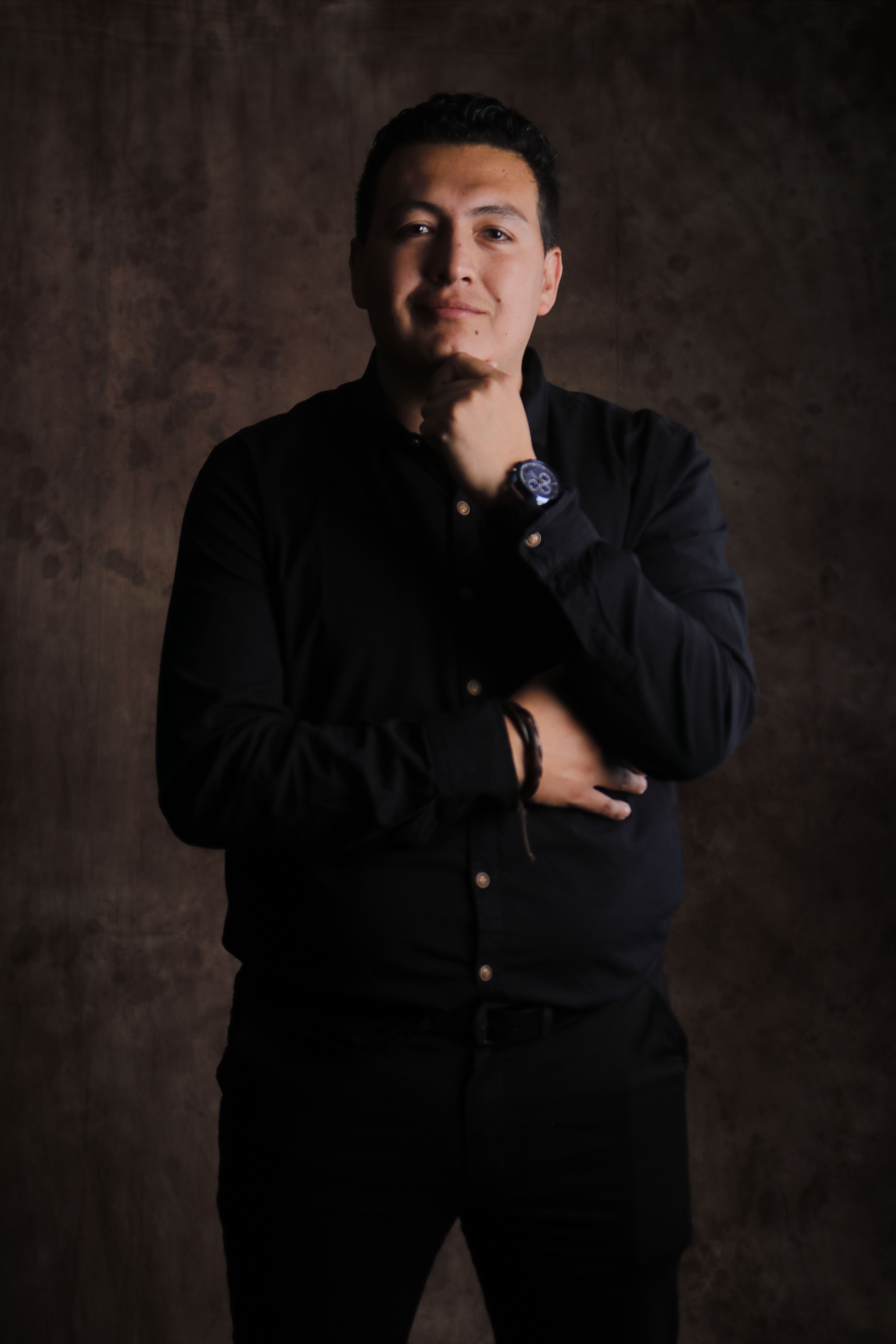 A man in dark clothing poses confidently against a textured brown background.