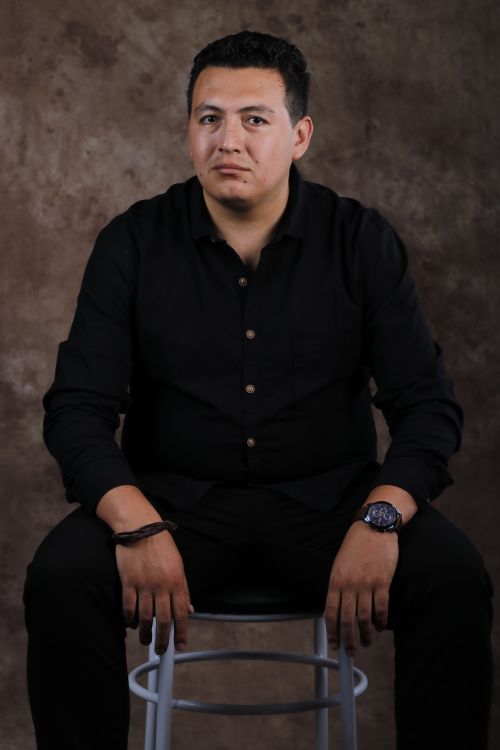 Free Portrait of a man in black attire seated on a stool against a brown backdrop. Stock Photo