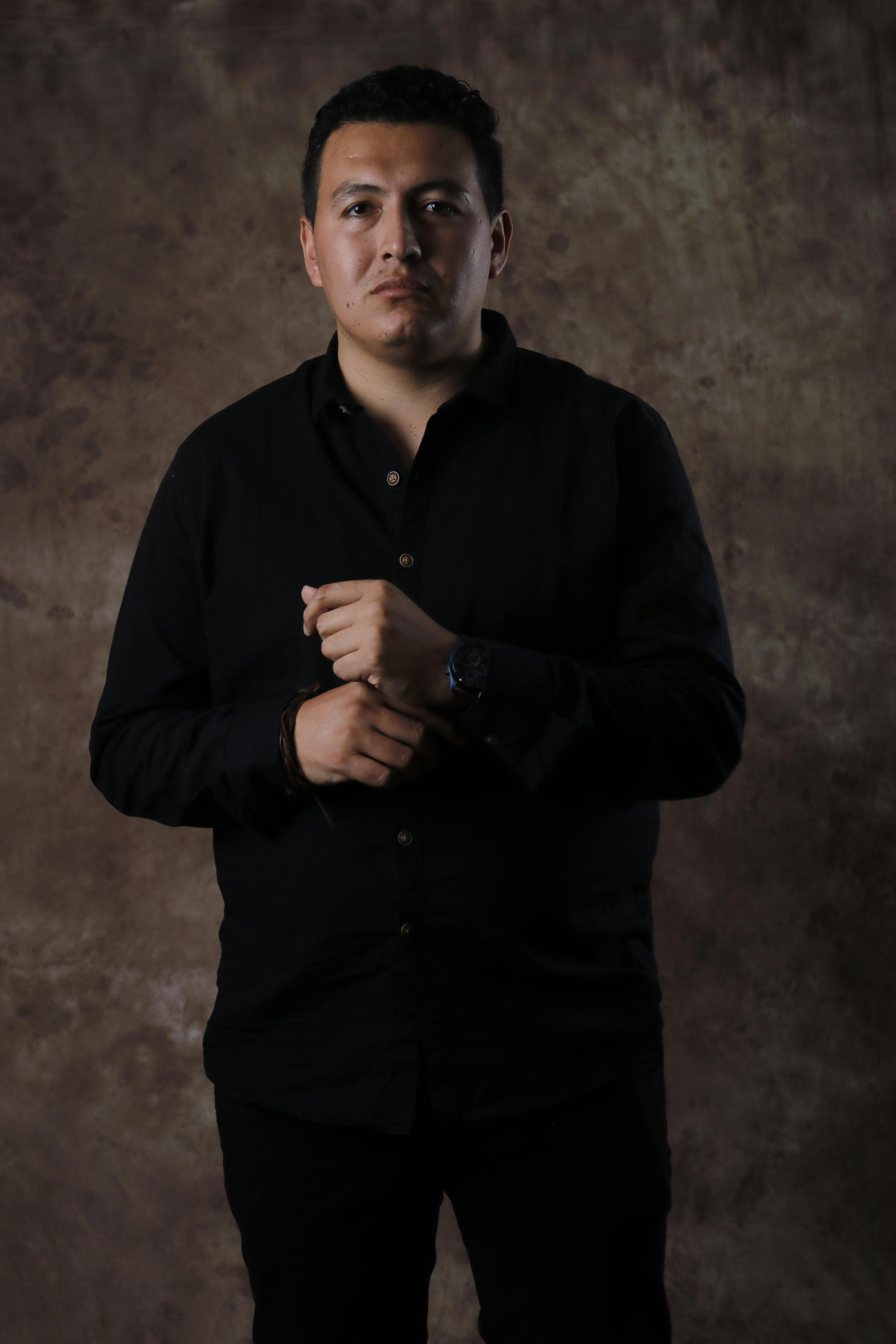 A man wearing a dark shirt poses against a textured brown backdrop in dramatic lighting.