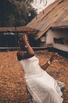 A woman in a white dress poses elegantly outdoors with a rustic backdrop.