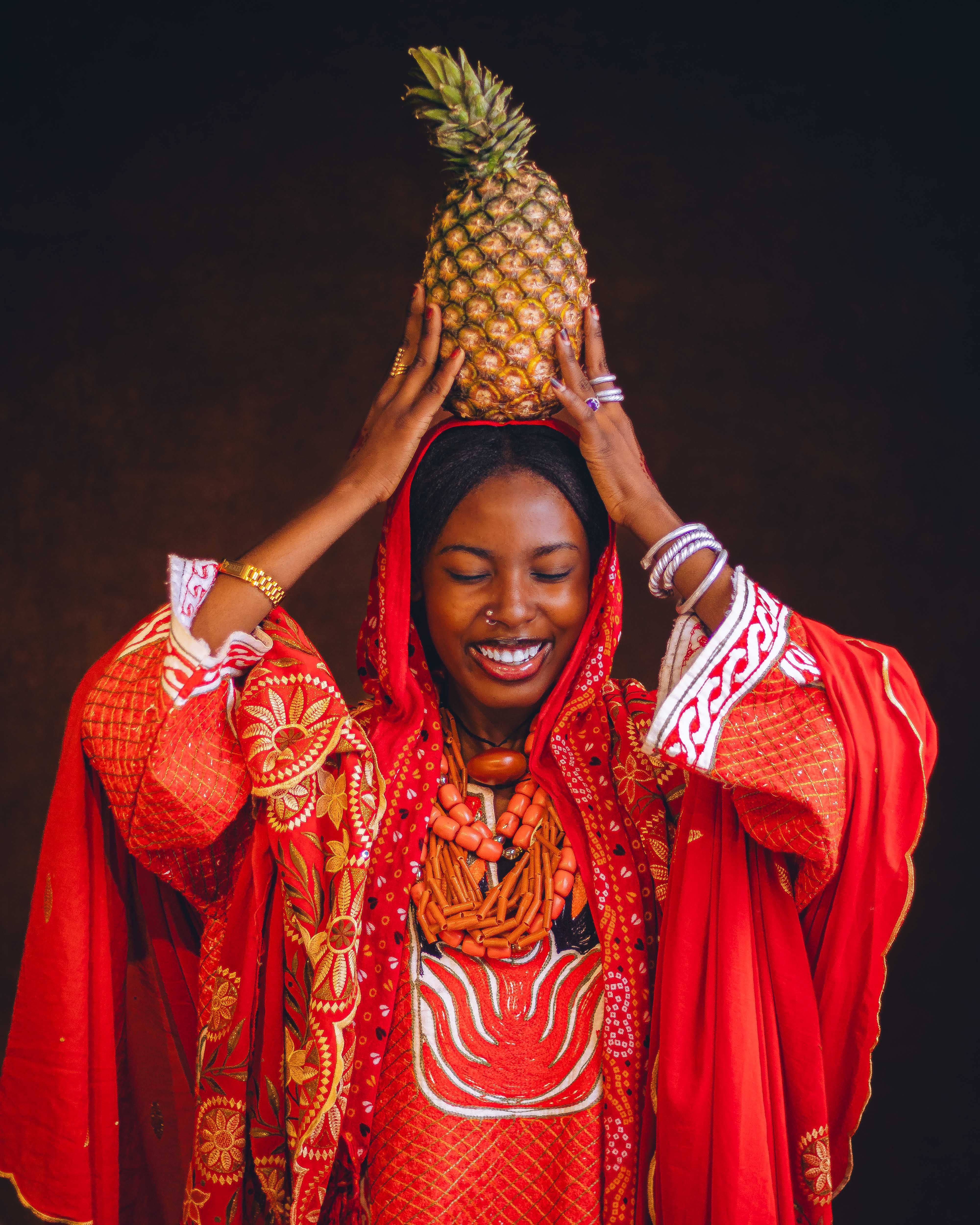 A smiling African woman in vibrant traditional attire joyfully holds a pineapple on her head, showcasing cultural heritage.