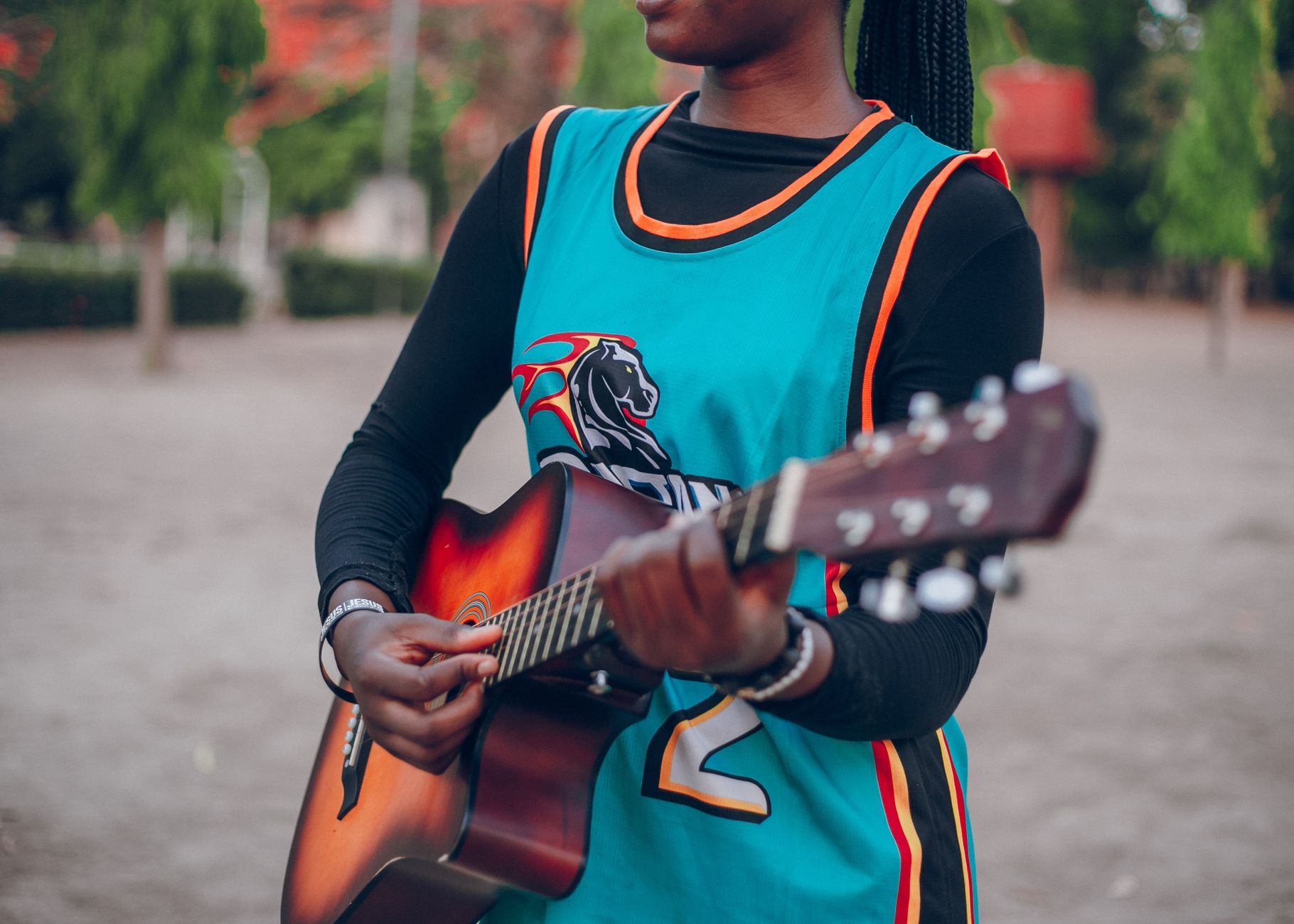 A person plays an acoustic guitar outdoors, wearing a sports jersey.