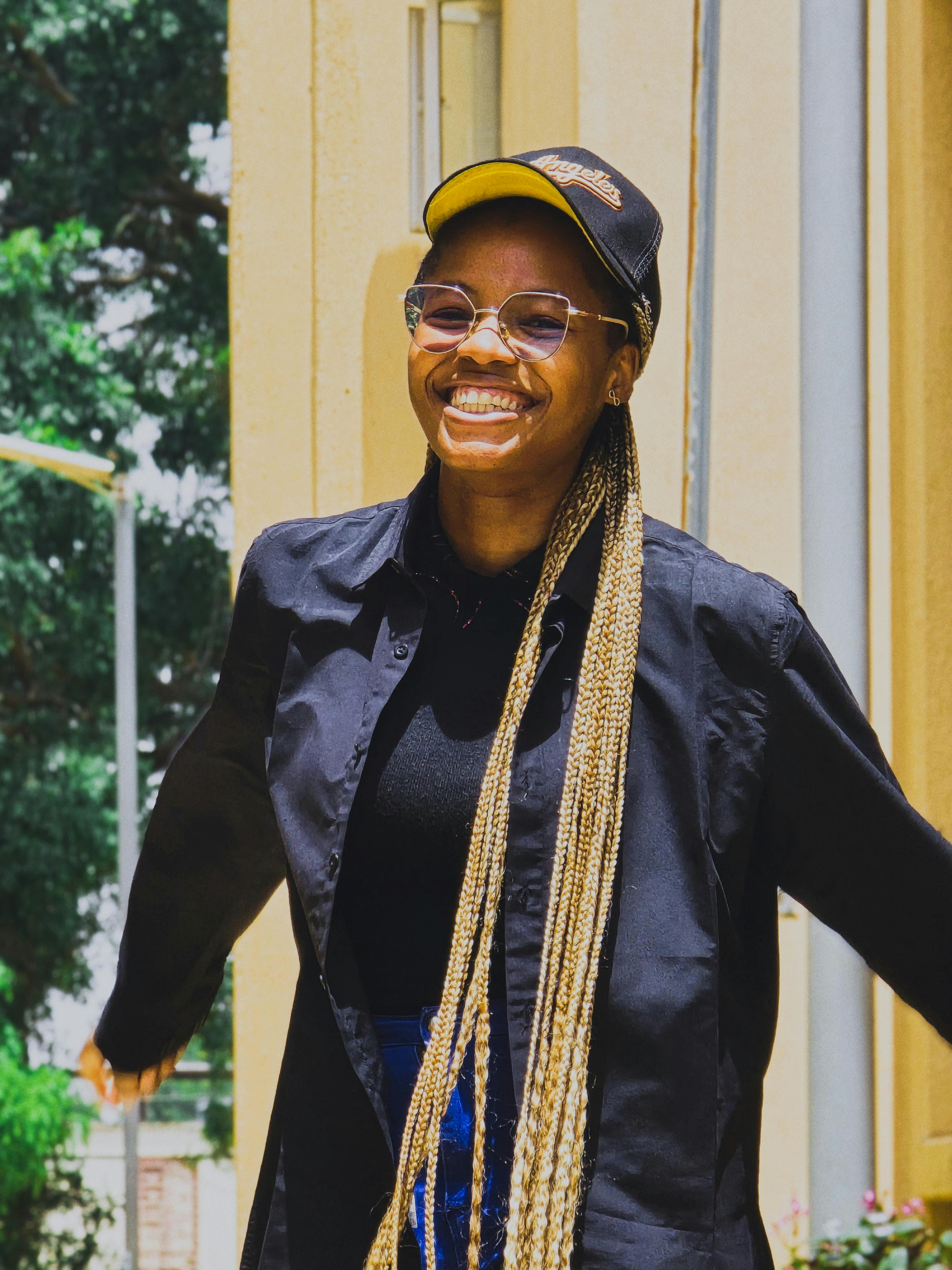 Joyful woman with long braids and cap, smiling outdoors in sunny weather.