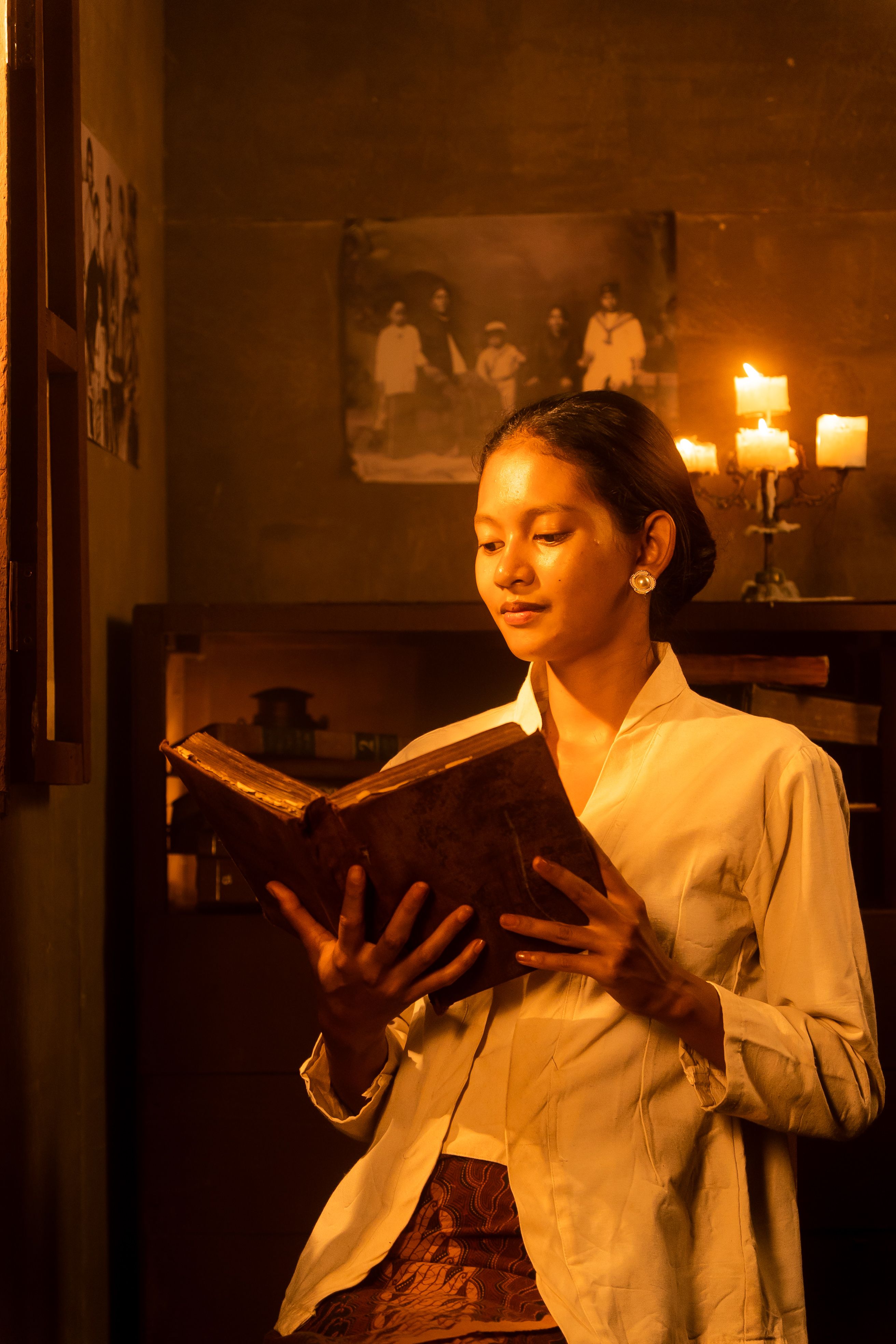 A young woman reads an old book in a vintage setting with candlelight.