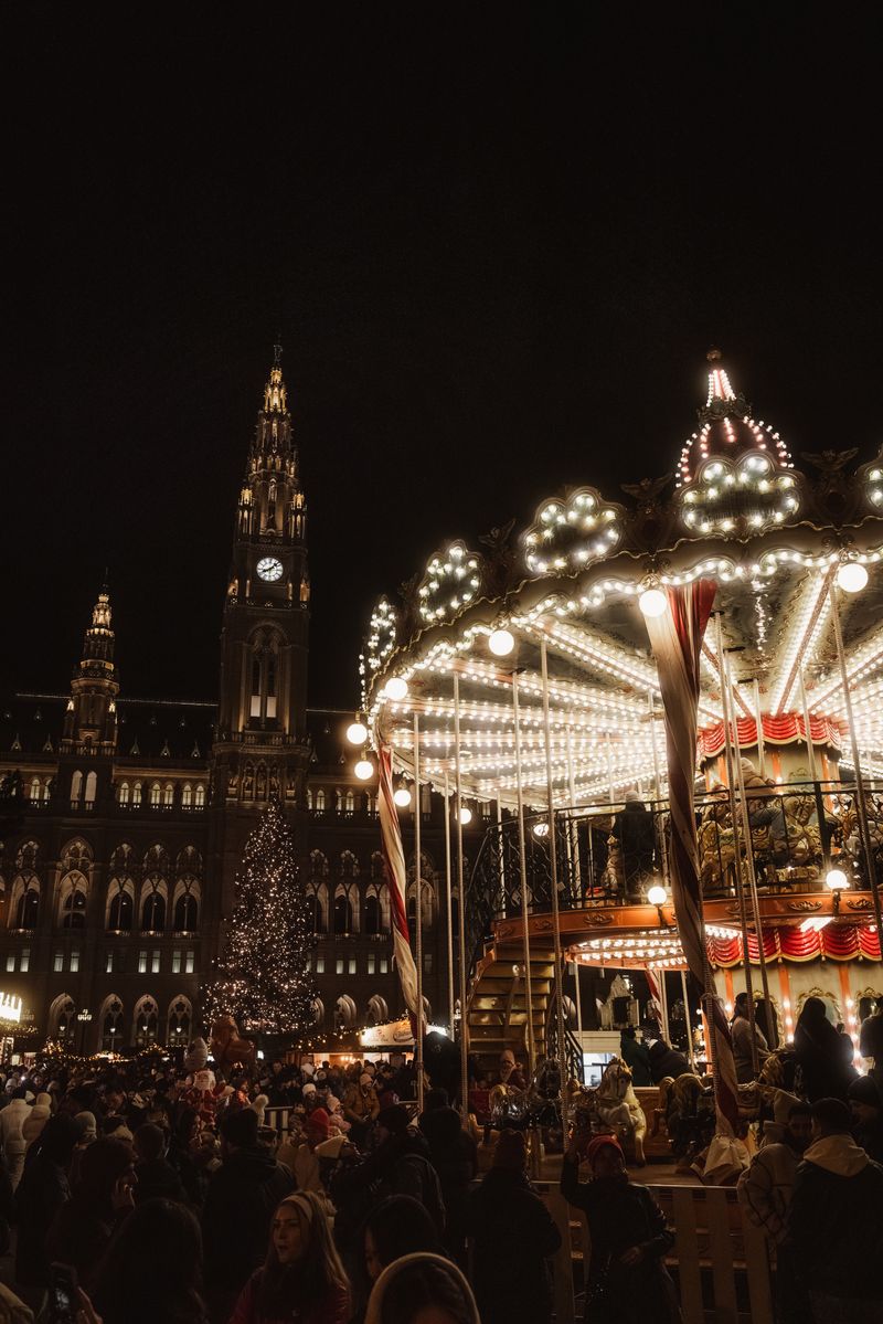 Bustling Christmas market scene at night with illuminated Rathaus and carousel in Vienna, Austria.